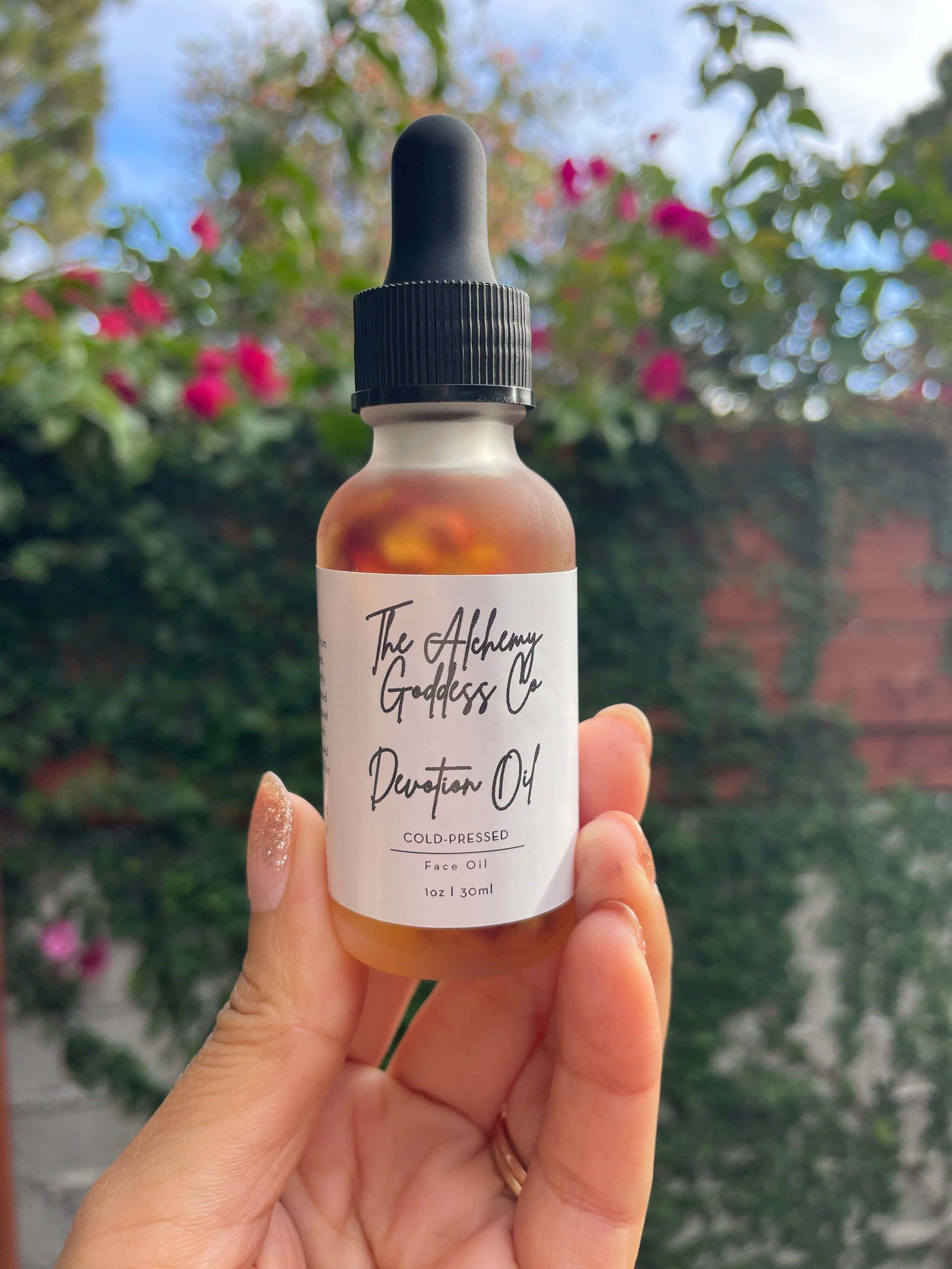 Rose Oil 桌布 壁紙 — 一隻手在明亮日光下，以綠色樹葉和粉紅色花為背景，握住一個標籤為「Devotion Oil」的琥珀色臉部護理油瓶。