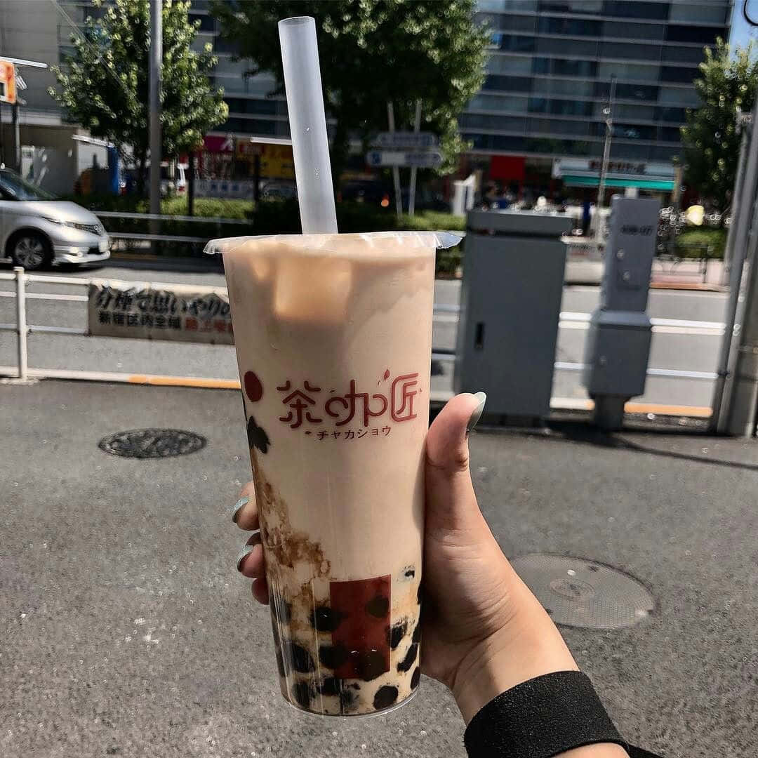 Boba Tea 桌布 壁紙 — 一隻手在明亮的城市街道背景下，握著一杯有黑色珍珠的奶油泡泡茶。