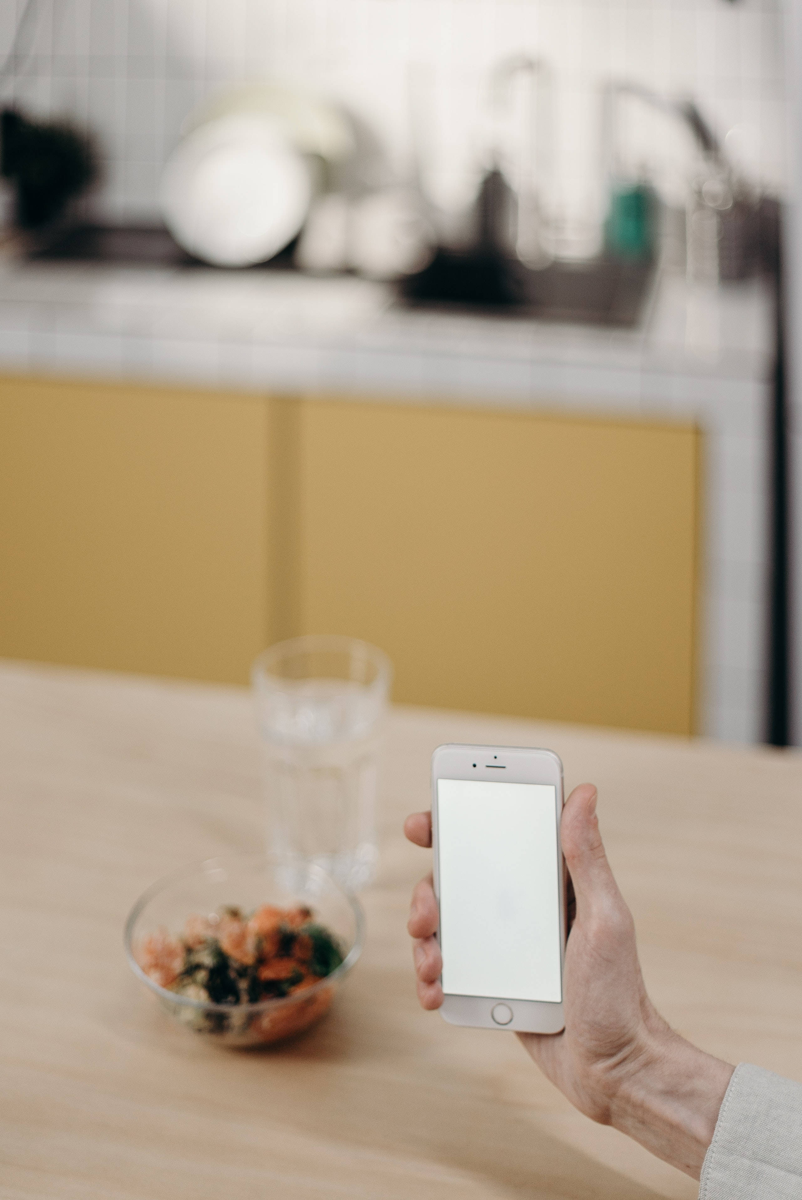 iPhone 桌布 壁紙 — 一隻手在明亮的廚房裡，拿著一部空白螢幕的智慧型手機，放在木桌上，旁邊有一碗沙拉和一杯水。