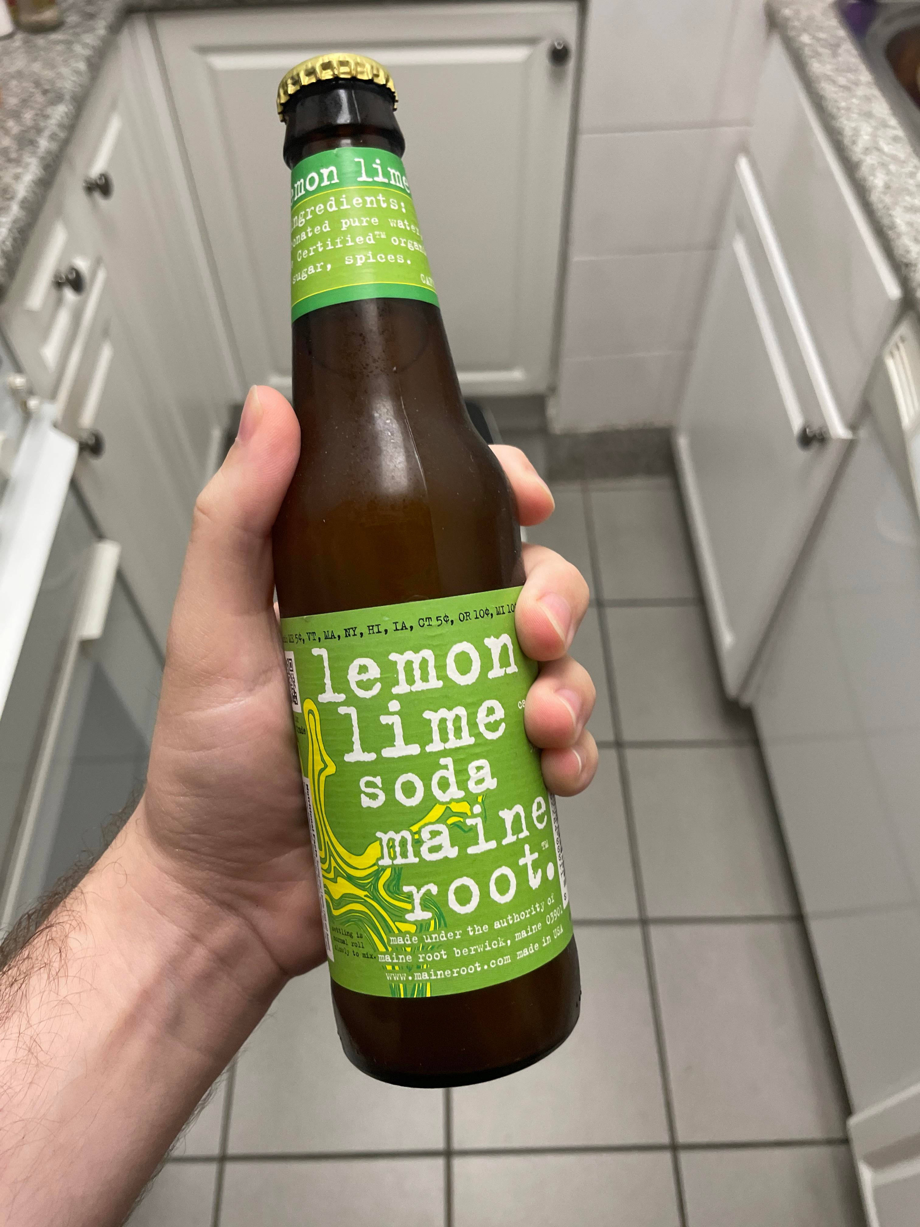 Drink 桌布 壁紙 — 一隻手在明亮的廚房裡拿著一個綠色標籤的Maine Root檸檬萊姆汽水瓶，廚房有白色櫥櫃和瓷磚地板。