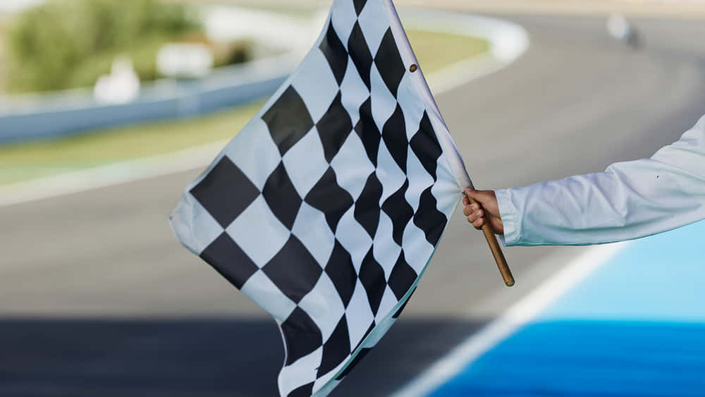 Chequered Flag 桌布 壁紙 — 一隻手在明亮的日光下，在彎曲的賽車道上揮舞著格子旗。