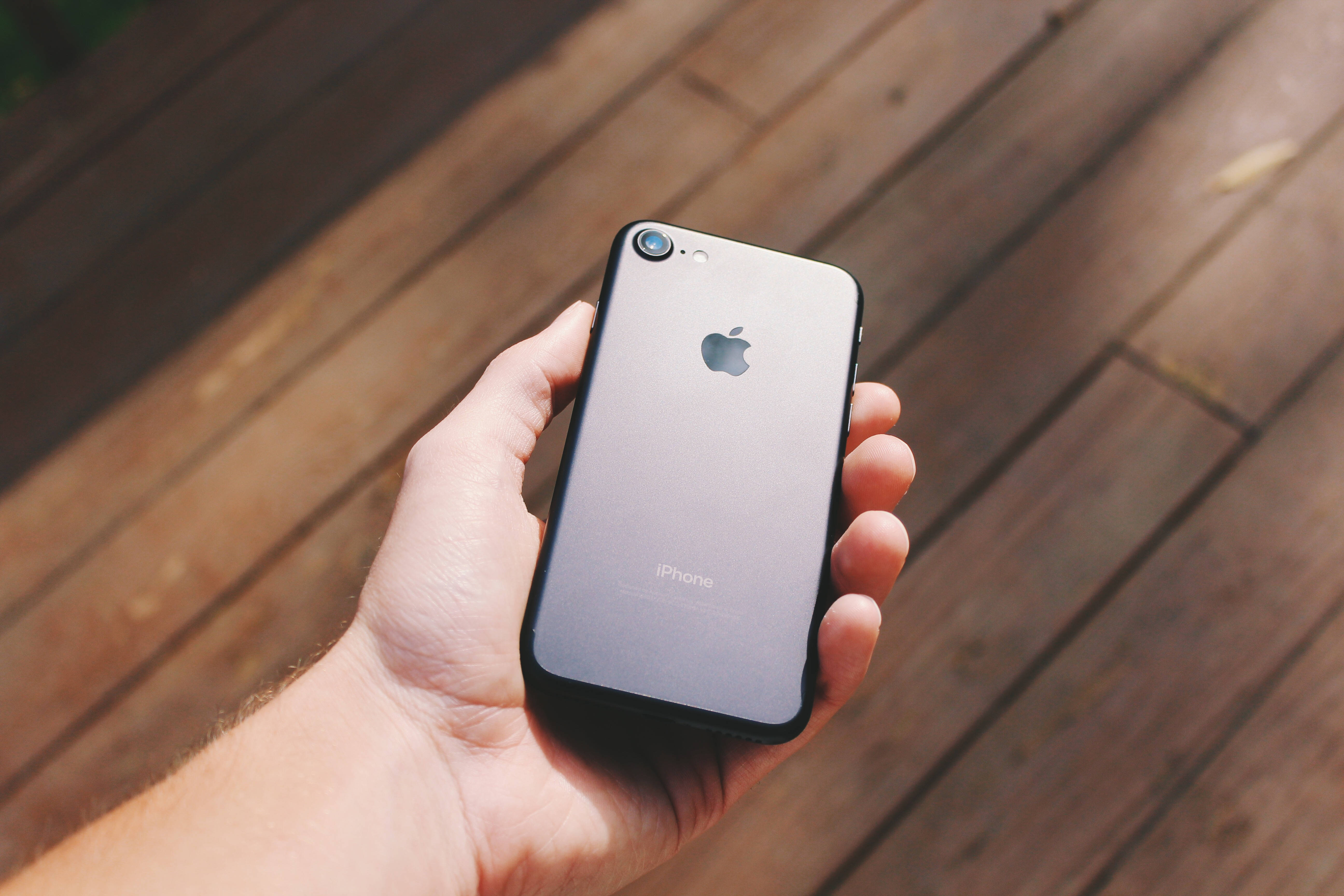 iPhone 8 桌布 壁紙 — 一隻手在明亮的陽光下，在溫暖的木質甲板上拿著銀色iPhone 7。