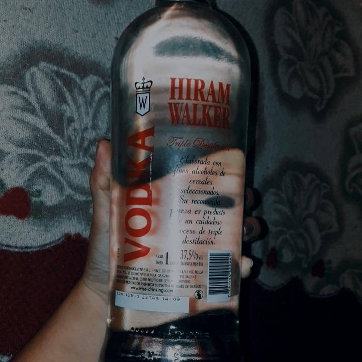 alcohol 桌布 壁紙 — 一隻手在昏暗燈光下，拿著Hiram Walker伏特加酒瓶，背景是有圖案的布料。