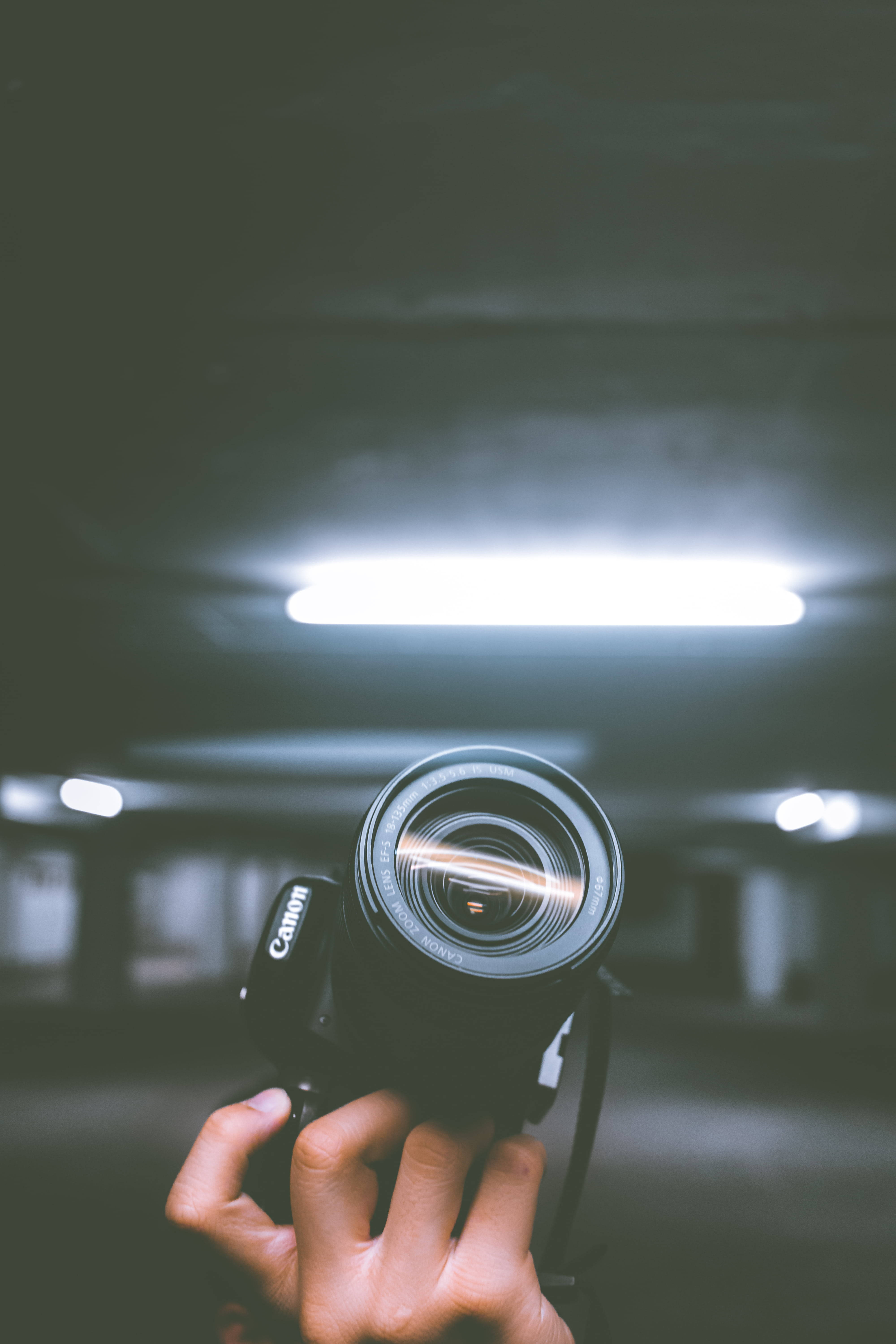 Light 桌布 壁紙 — 一隻手在昏暗的地下停車場中，於明亮的螢光燈下拿著一部黑色Canon相機。