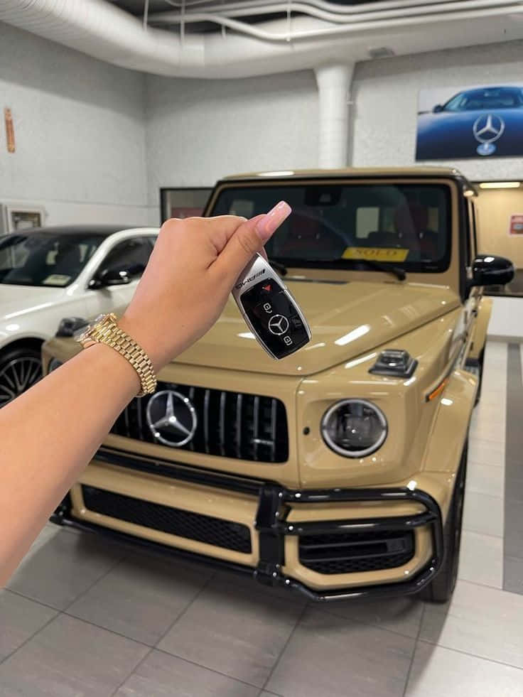 Off-road Car 桌布 壁紙 — 一隻手在時尚的汽車經銷商展廳中，拿著一個賓士車鑰匙遙控器，前面是一輛米色G-Wagen。