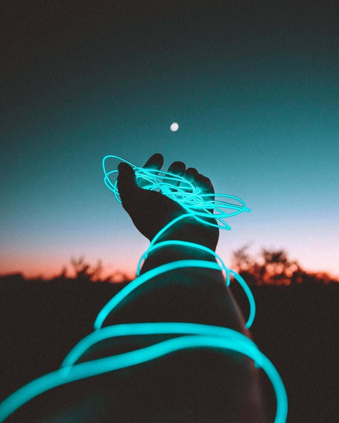 Light 桌布 壁紙 — 一隻手在暮色天空中握著發光的青色光絲，背景有可見的月亮和剪影樹木。