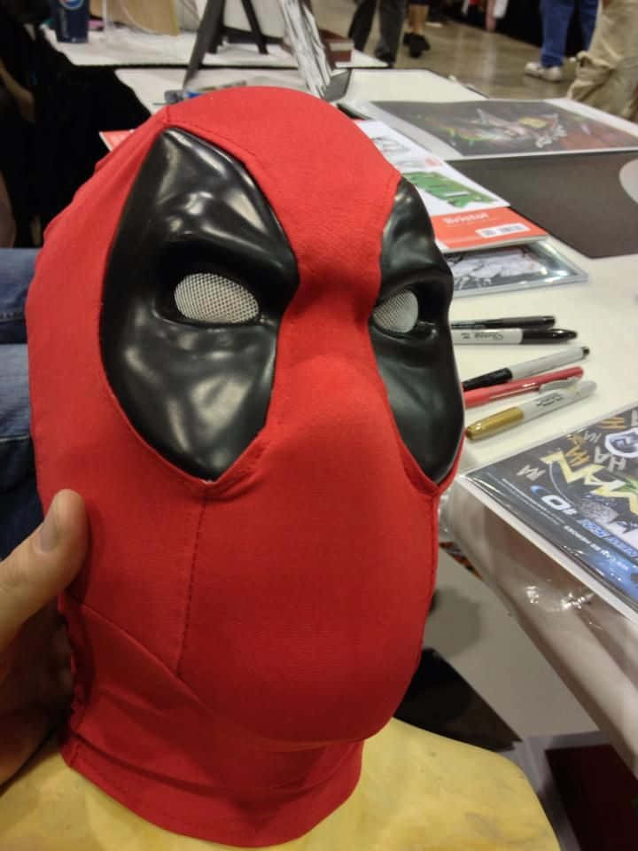 Deadpool Mask 桌布 壁紙 — 一隻手在會議桌上拿著一個帶有網眼眼孔的紅黑色死侍面具。