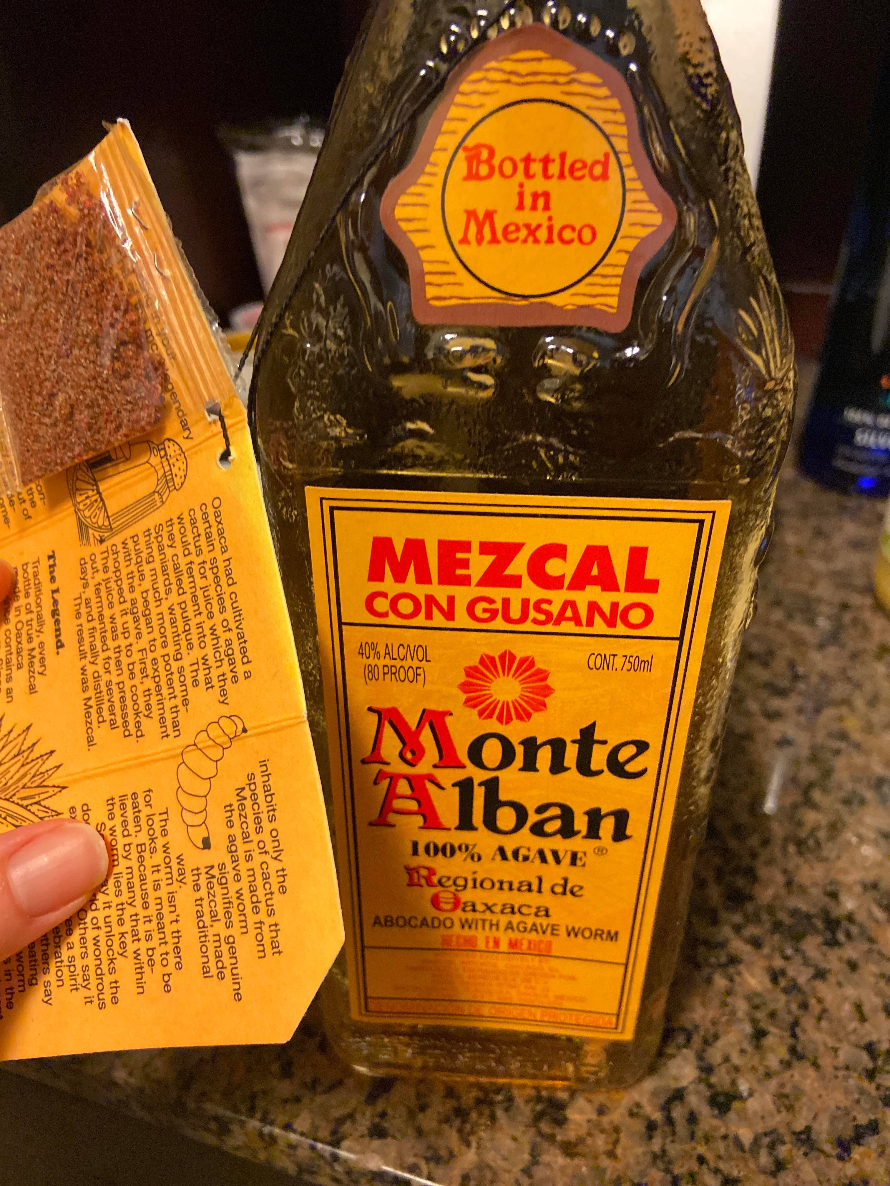 Drink 桌布 壁紙 — 一隻手在有斑點的檯面上，在一瓶Monte Alban梅斯卡爾酒旁邊拿著一包梅斯卡爾蟲。