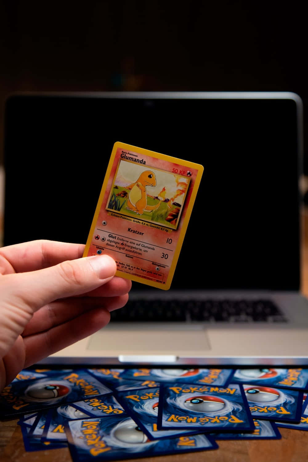 Pokemon Card 桌布 壁紙 — 一隻手在木桌上拿著一張復古Glumanda Pokémon卡，在筆記型電腦上方，周圍散落著精靈球卡。