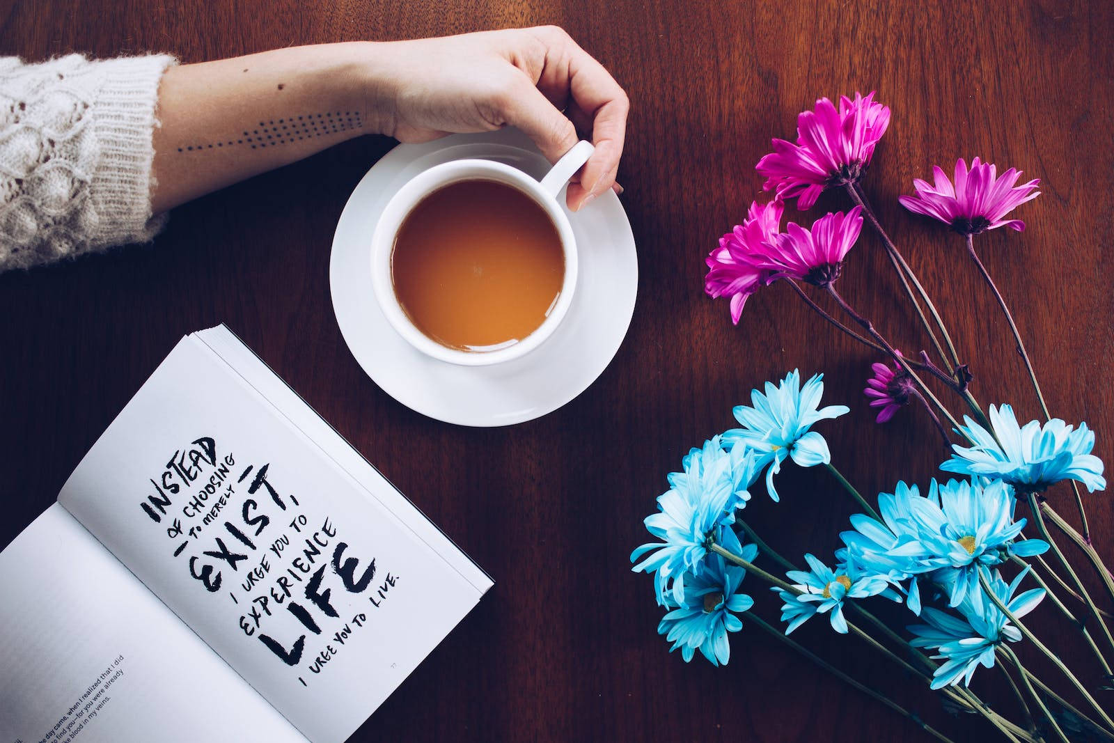 Motivational Quotes 桌布 壁紙 — 一隻手在木桌上拿著一杯茶，旁邊有一本打開的書和鮮豔的粉紅色和藍色花朵。