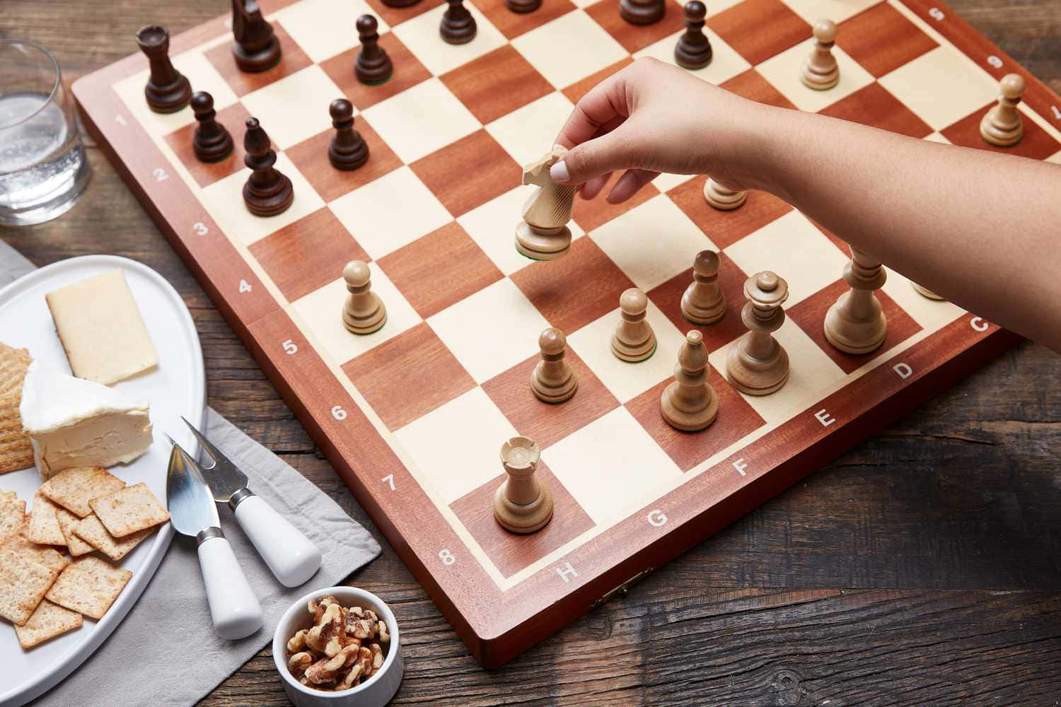 Chessboard 桌布 壁紙 — 一隻手在木製棋盤上移動西洋棋兵，旁邊放著一盤起司餅乾和堅果，置於鄉村風格的桌子上。