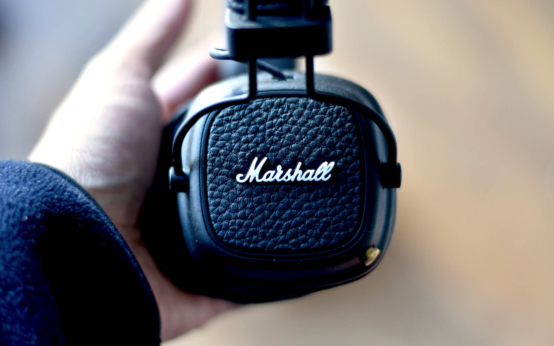 Marshall 桌布 壁紙 — 一隻手在柔和焦點背景中，拿著一個黑色Marshall頭戴式耳機，耳罩有紋理皮革和白色草書標誌。