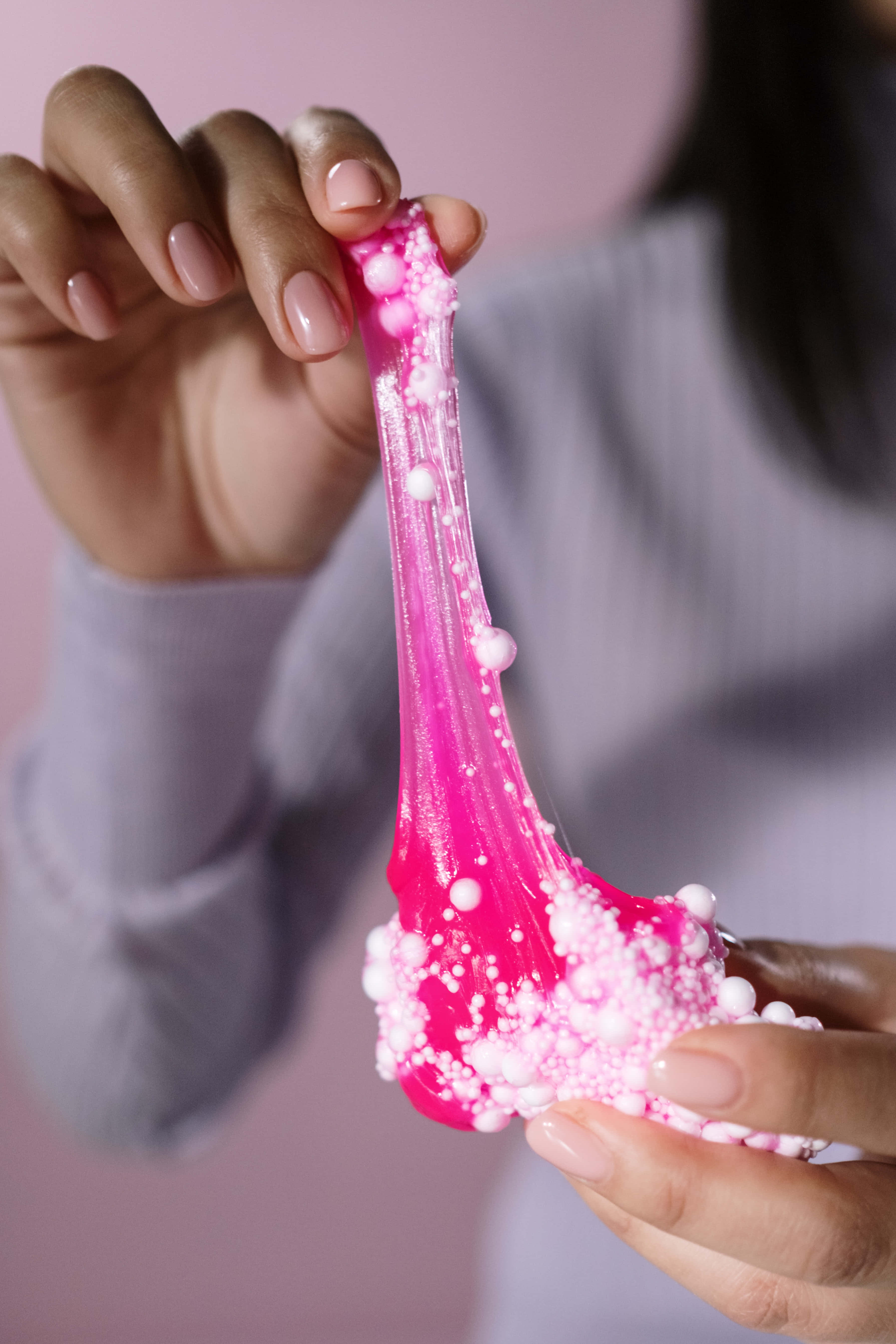 slime 桌布 壁紙 — 一隻手在柔和的粉紅色背景上拉伸充滿活力的粉紅色閃粉史萊姆，上面點綴著白色珠子。