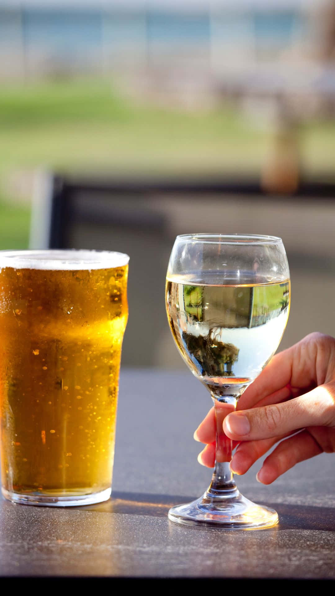 Beer 桌布 壁紙 — 一隻手在模糊的戶外背景下，拿著一個酒杯放在一個冒泡的啤酒杯旁邊的桌子上。