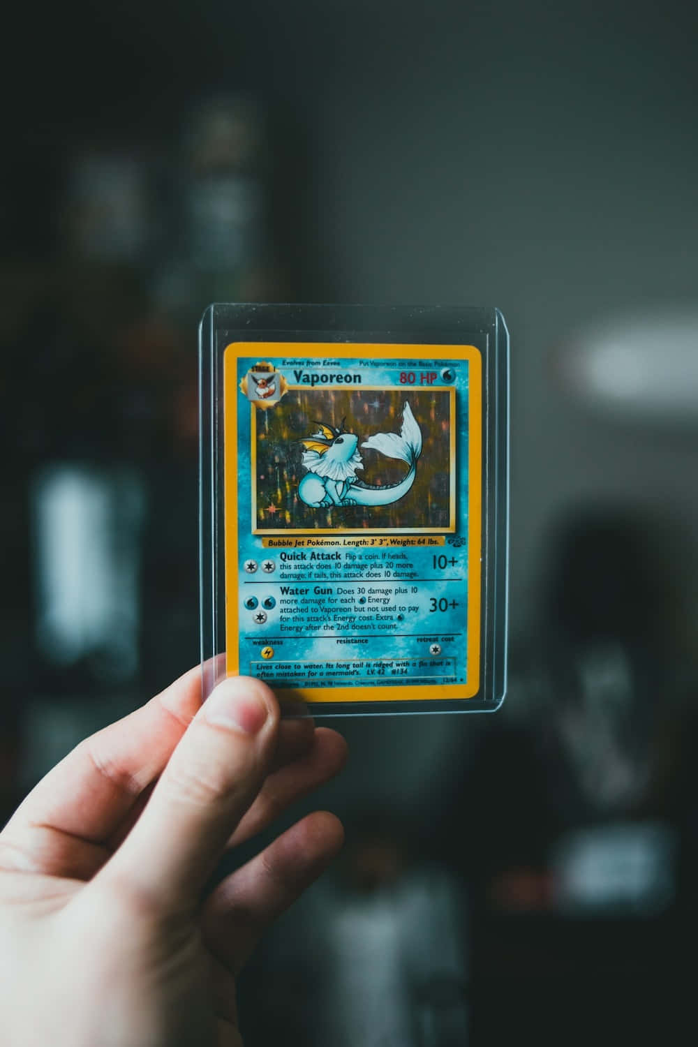 Pokemon Card 桌布 壁紙 — 一隻手在模糊的深色背景前，拿著一張裝在透明保護套中的復古 Vaporeon 寶可夢卡。
