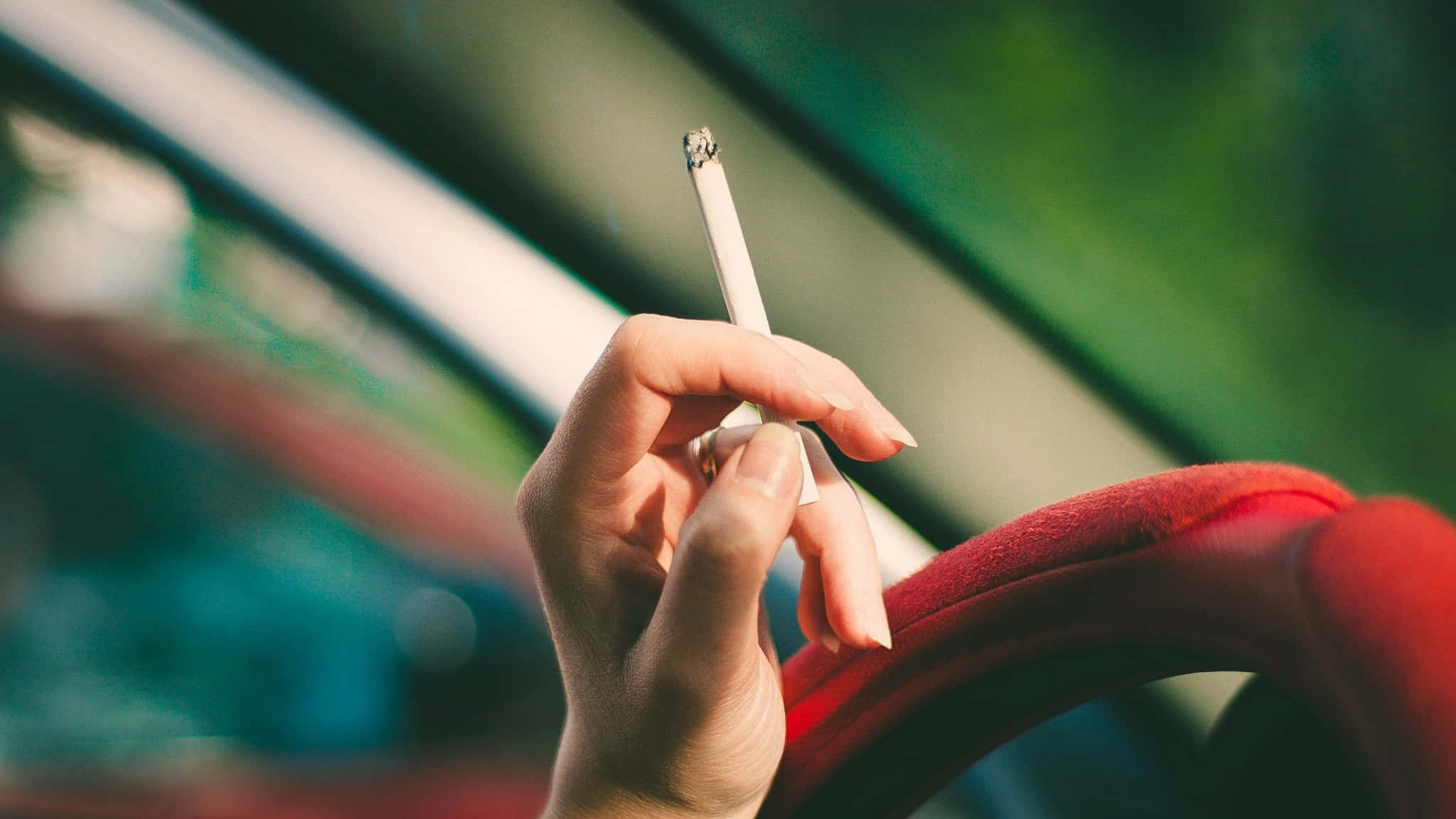 driving 桌布 壁紙 — 一隻手在模糊的車廂內拿著一支點燃的香煙，車廂有紅色的座椅，外面有綠色的樹葉。