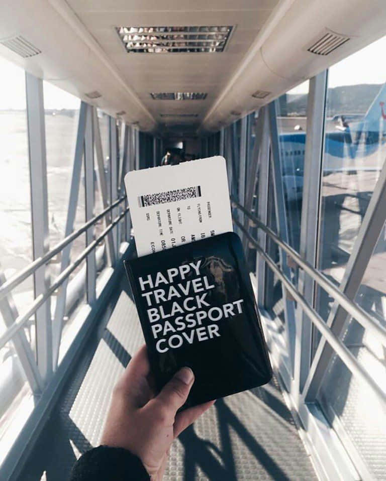 Passport 桌布 壁紙 — 一隻手在機場登機橋上，在明亮的陽光下拿著一個帶有白色文字的黑色護照套和登機證。