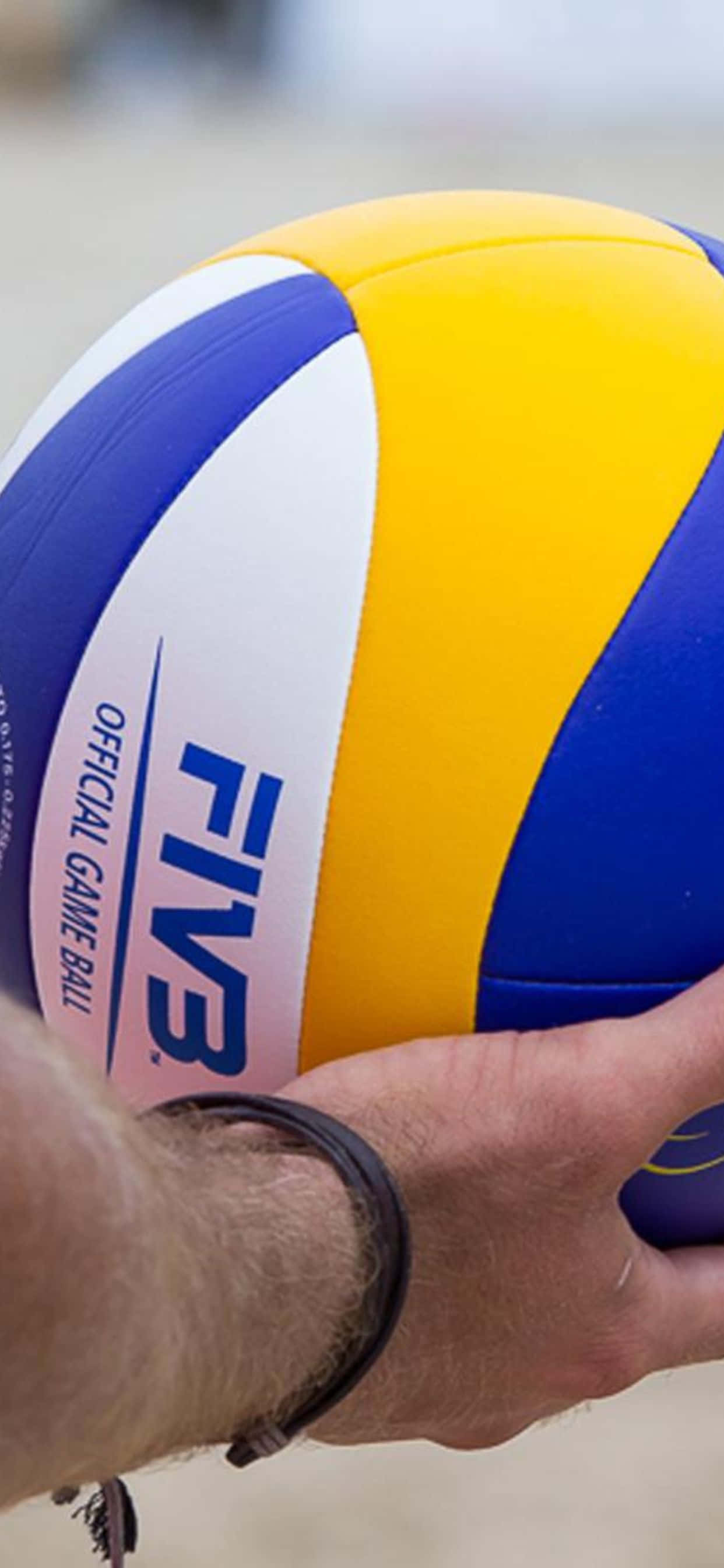 iPhone Xs Max Volleyball 桌布 壁紙 — 一隻手在沙灘上緊握著鮮豔的藍、黃、白色FIVB官方排球的特寫。