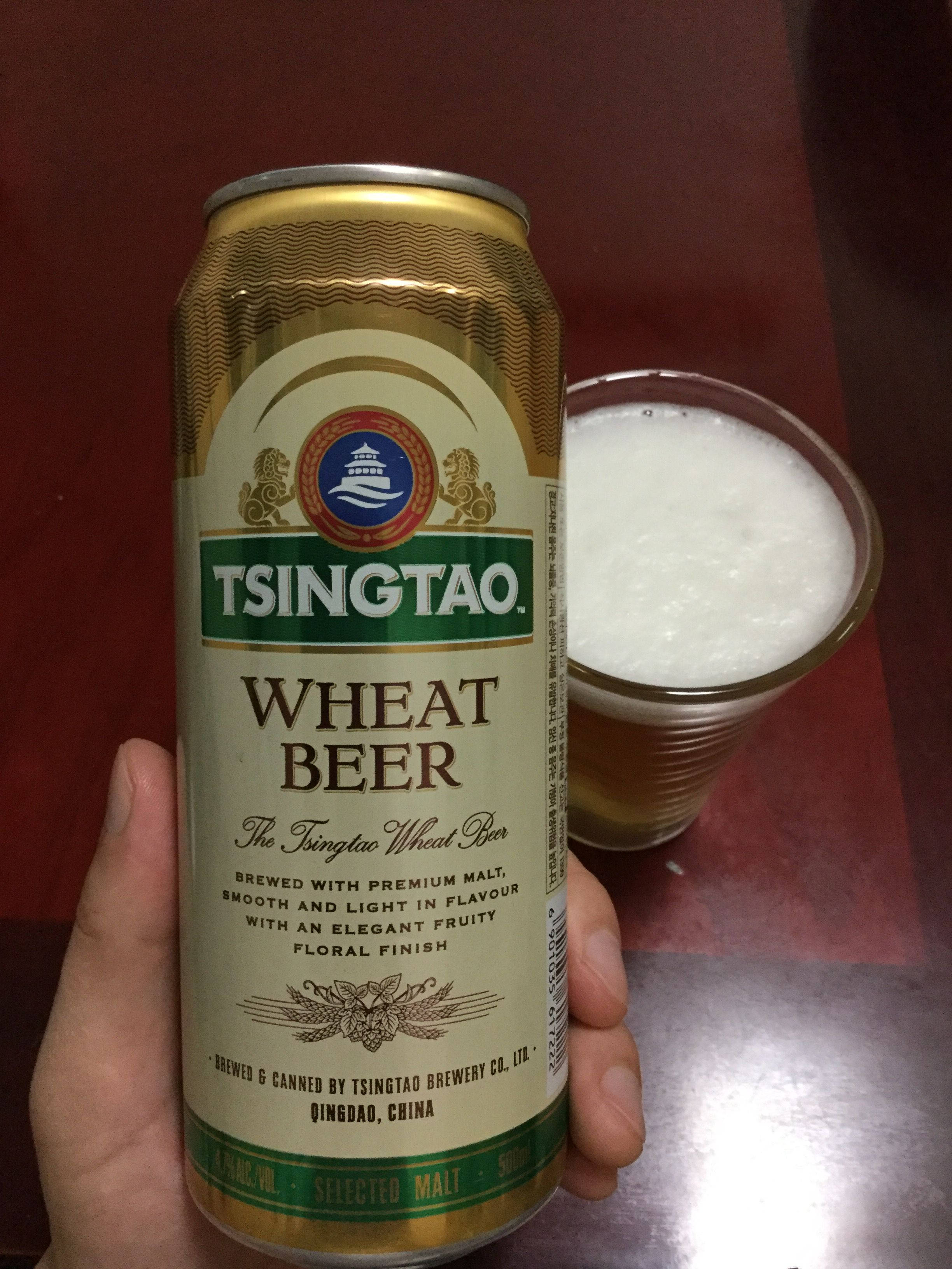 booze 桌布 壁紙 — 一隻手在深紅色表面上拿著一個金色青島小麥啤酒罐，旁邊放著一個裝滿泡沫啤酒的玻璃杯。