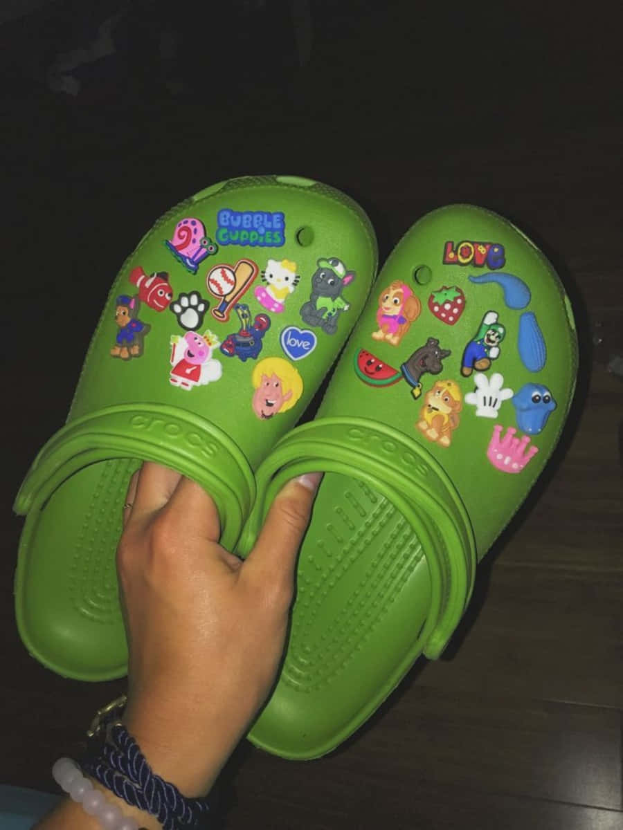 Crocs 桌布 壁紙 — 一隻手在深色木地板上拿著一雙鮮綠色Crocs，上面裝飾了色彩繽紛的卡通角色貼紙。