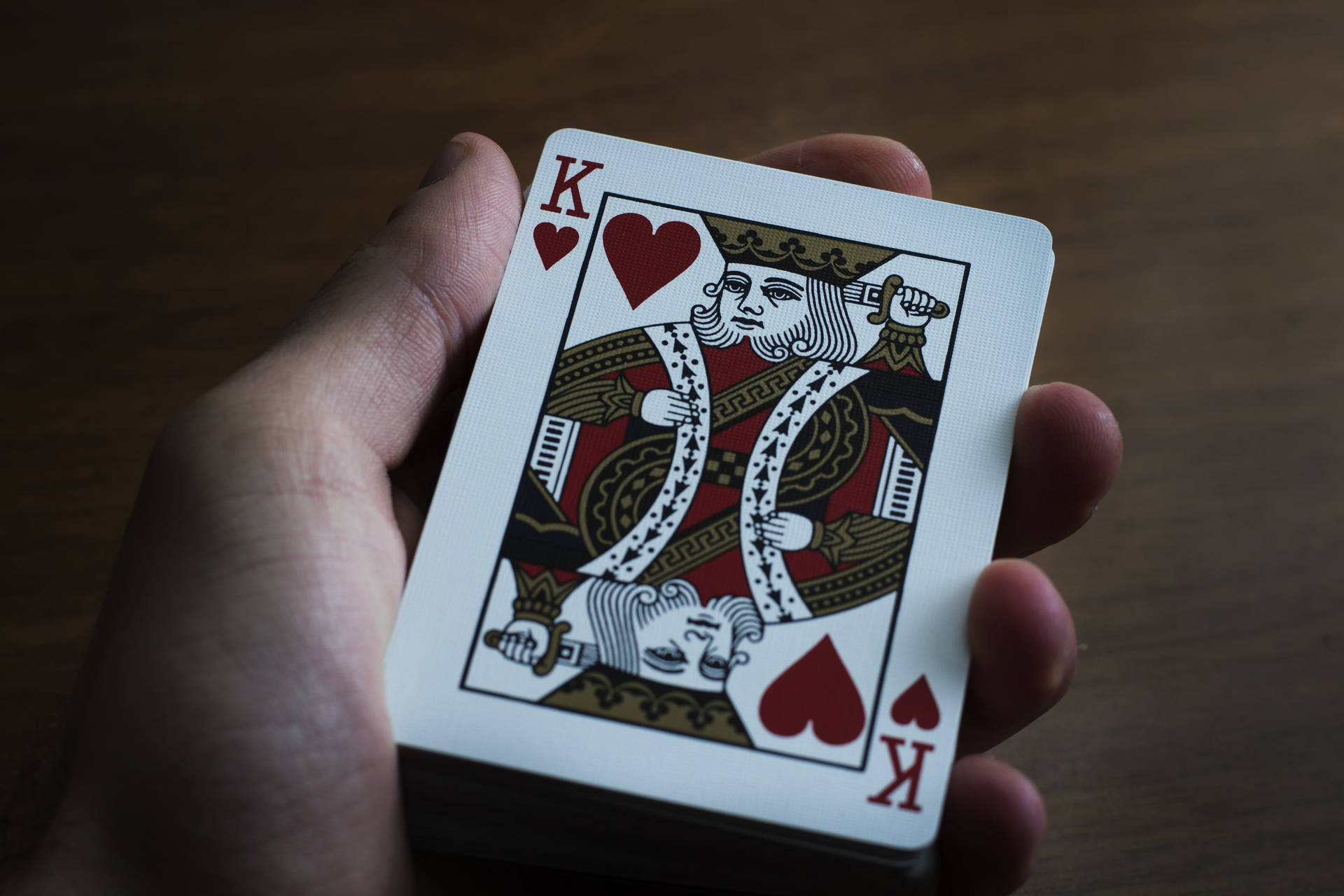 Playing Cards 桌布 壁紙 — 一隻手在深色木質表面上用柔和光線拿著一張紅心K撲克牌。
