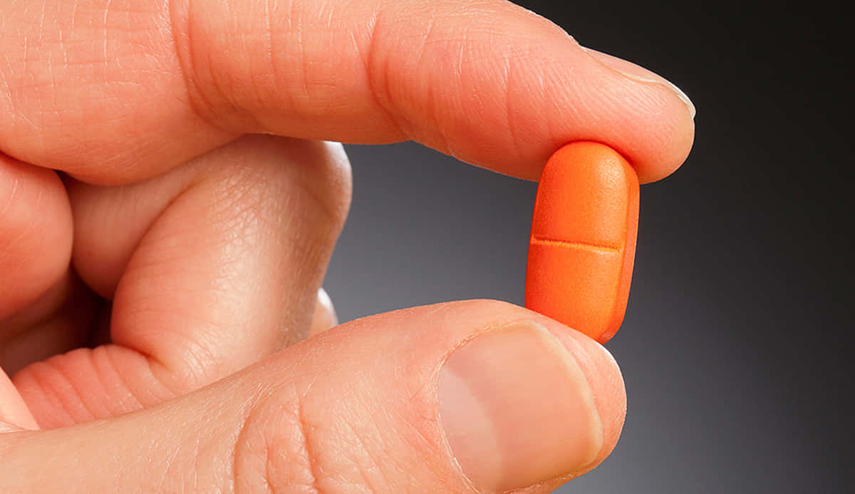 pills 桌布 壁紙 — 一隻手在深色漸變背景上輕輕拿著橙色藥丸的特寫。