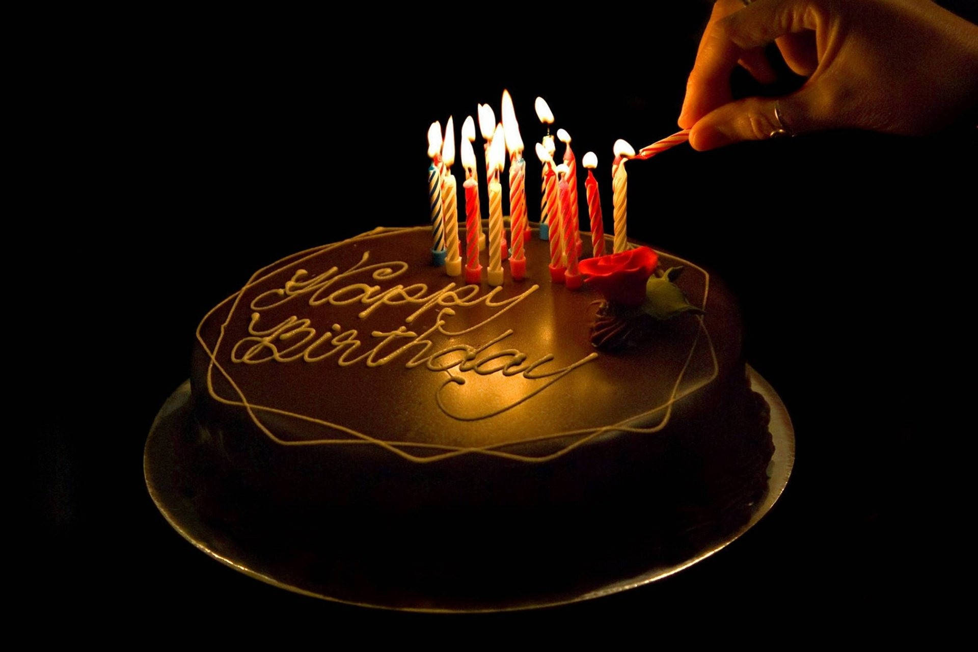 蛋糕 桌布 壁紙 — 一隻手在深色背景前，點燃一個帶有 'Happy Birthday' 糖霜的巧克力生日蛋糕上的蠟燭。