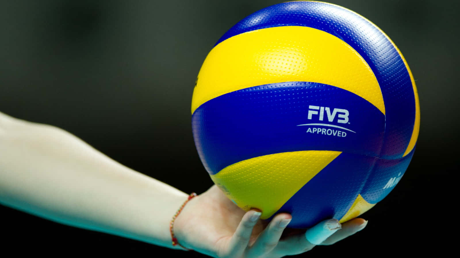 Volleyball Ball 桌布 壁紙 — 一隻手在深色背景前拿著一個藍黃相間、經FIVB認證的排球。