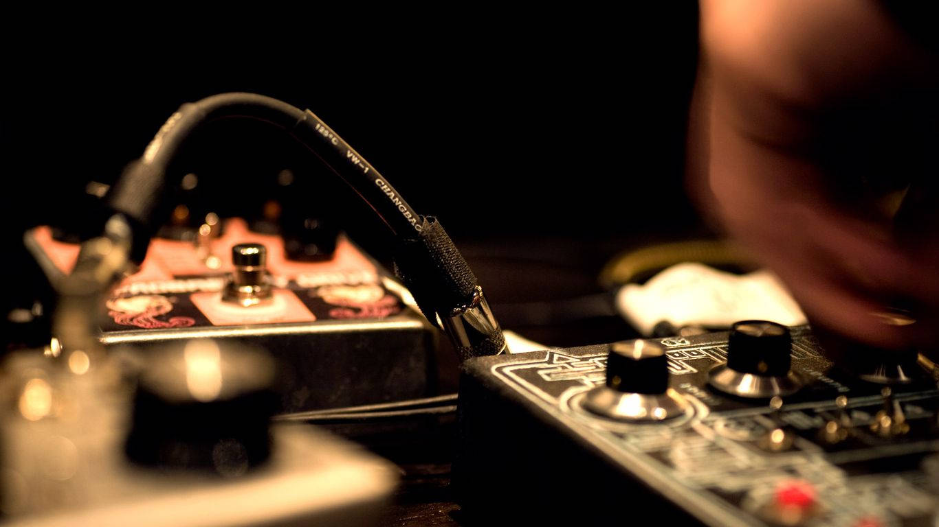 Beautiful Music 桌布 壁紙 — 一隻手在溫暖昏暗的燈光下調整一個帶有紅色指示燈的黑色吉他效果踏板的旋鈕。