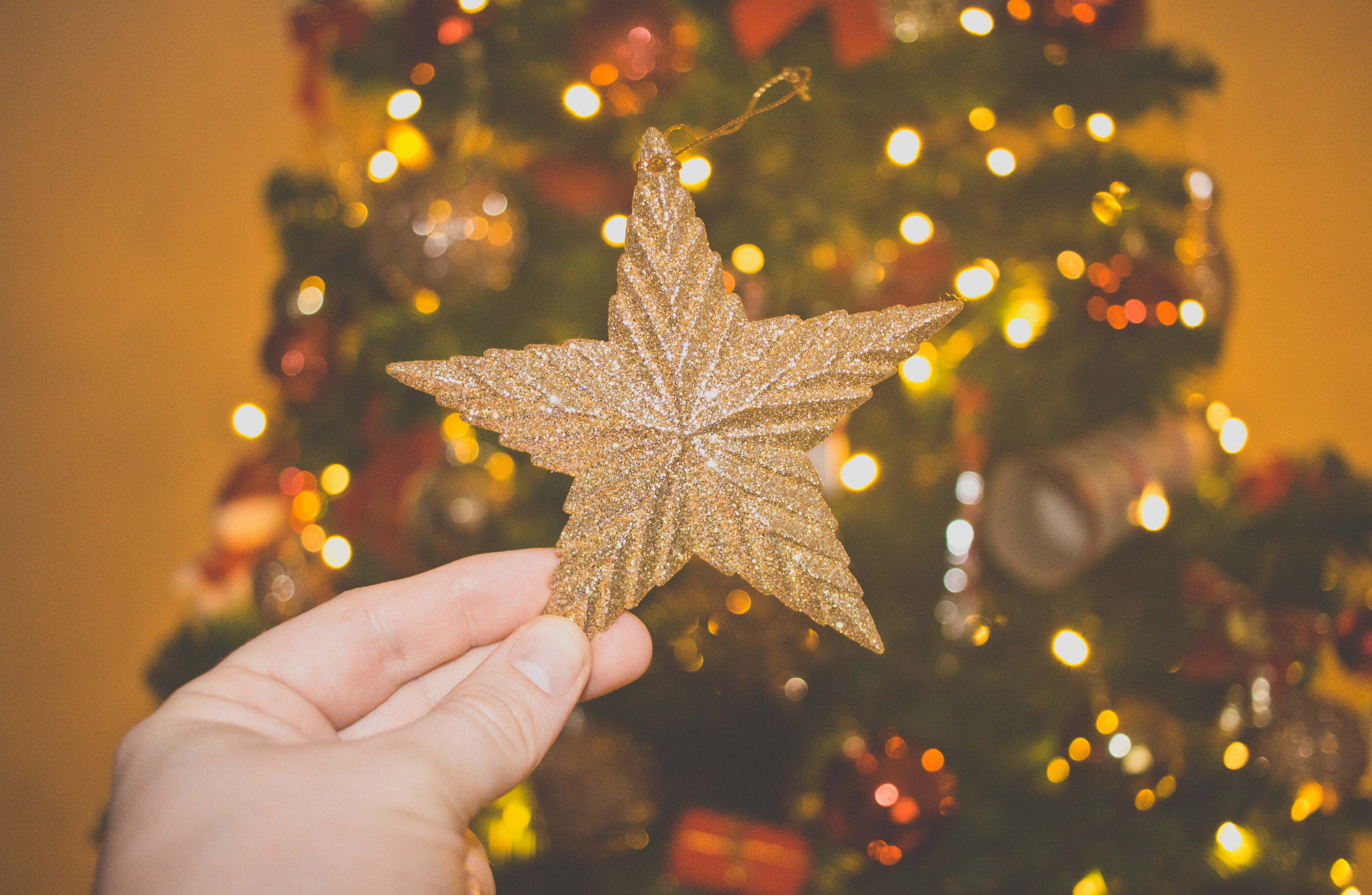 Christmas Star 桌布 壁紙 — 一隻手在溫暖燈光下模糊的聖誕樹前，拿著一個閃閃發光的金色星星裝飾。