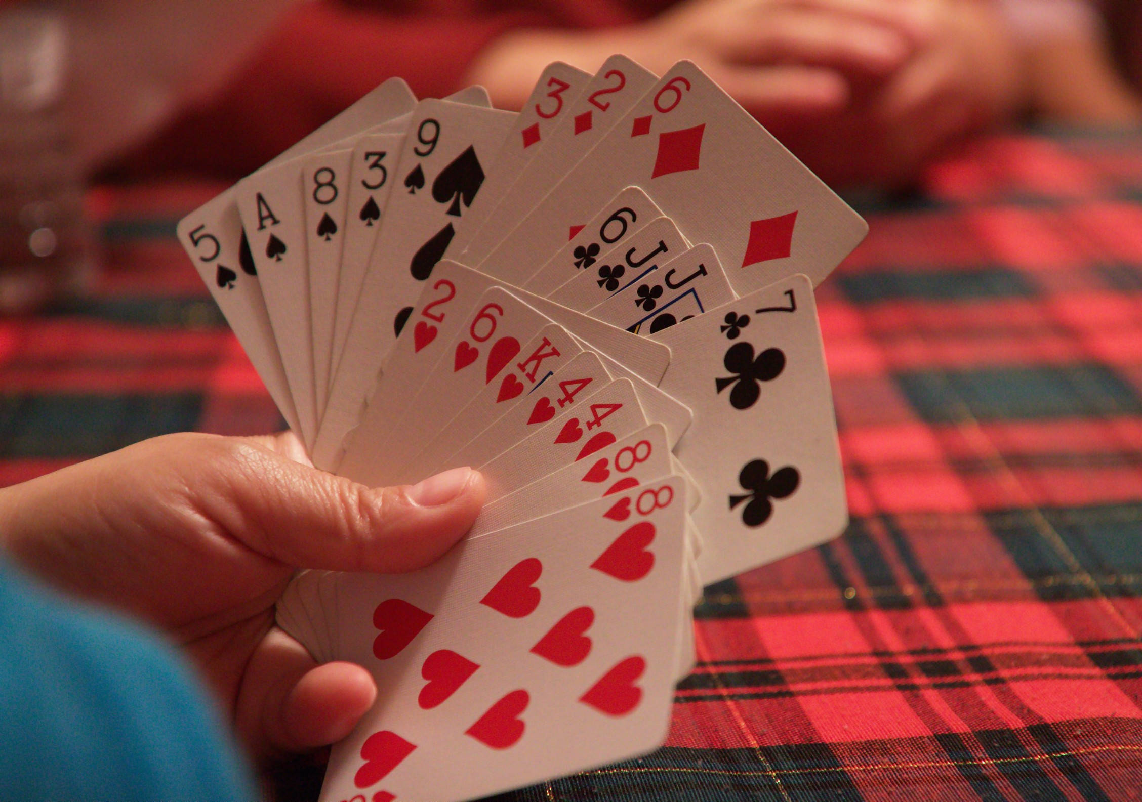 Playing Cards 桌布 壁紙 — 一隻手在溫暖的室內燈光下，將撲克牌扇開在紅色格子桌布上。