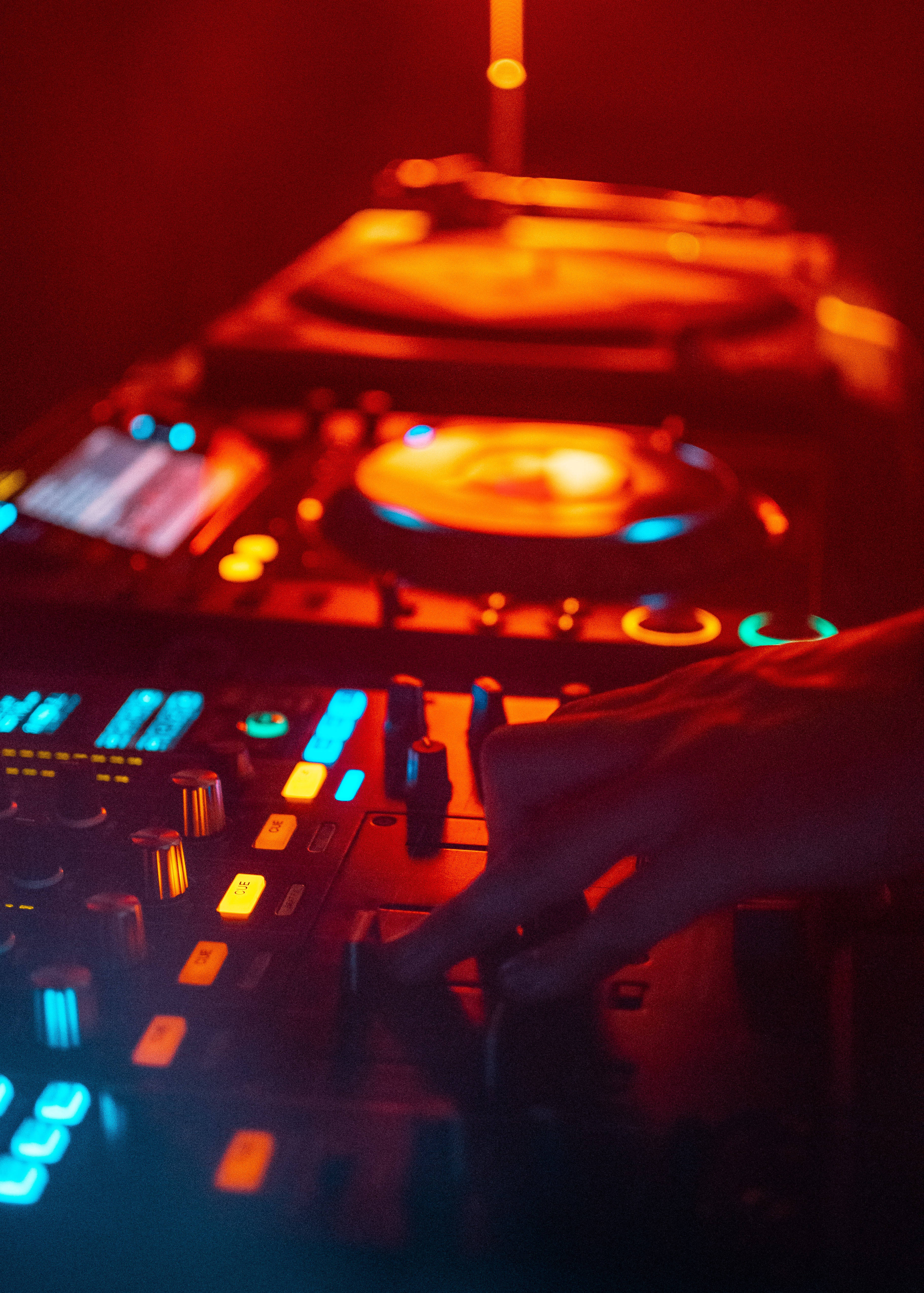 DJ 桌布 壁紙 — 一隻手在溫暖的紅燈下調整DJ混音器上發光的藍色旋鈕，背景中的唱盤模糊不清。
