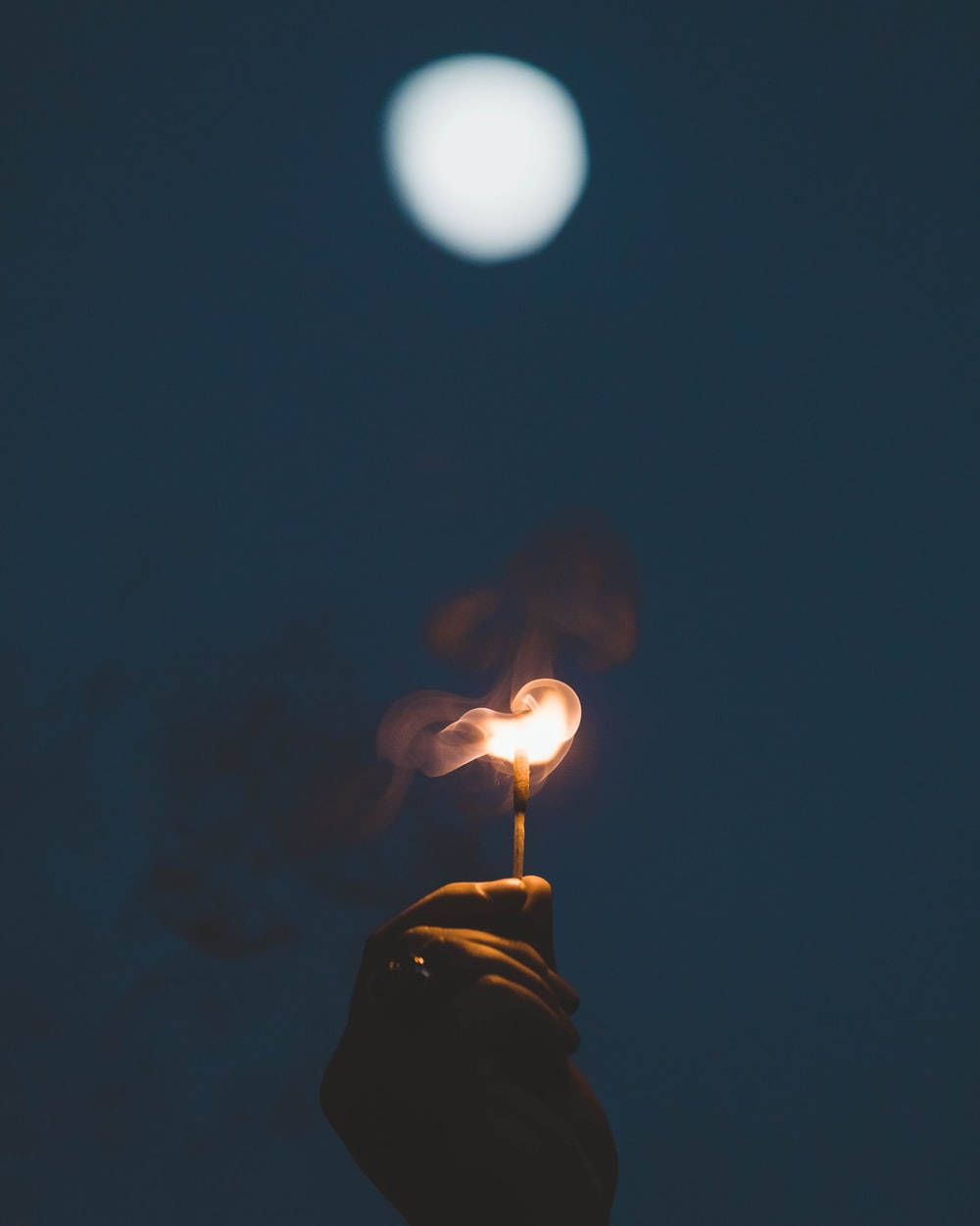火焰動畫 桌布 壁紙 — 一隻手在漆黑的夜空下拿著一根點燃的火柴，上方一輪發光的月亮將柔和的光線灑在火焰和煙霧上。