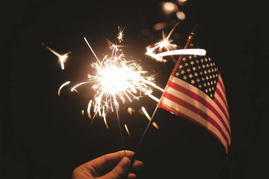 USA flag iPhone 桌布 壁紙 — 一隻手在漆黑的夜空下拿著閃爍的煙火和美國國旗，周圍有發光的火星。