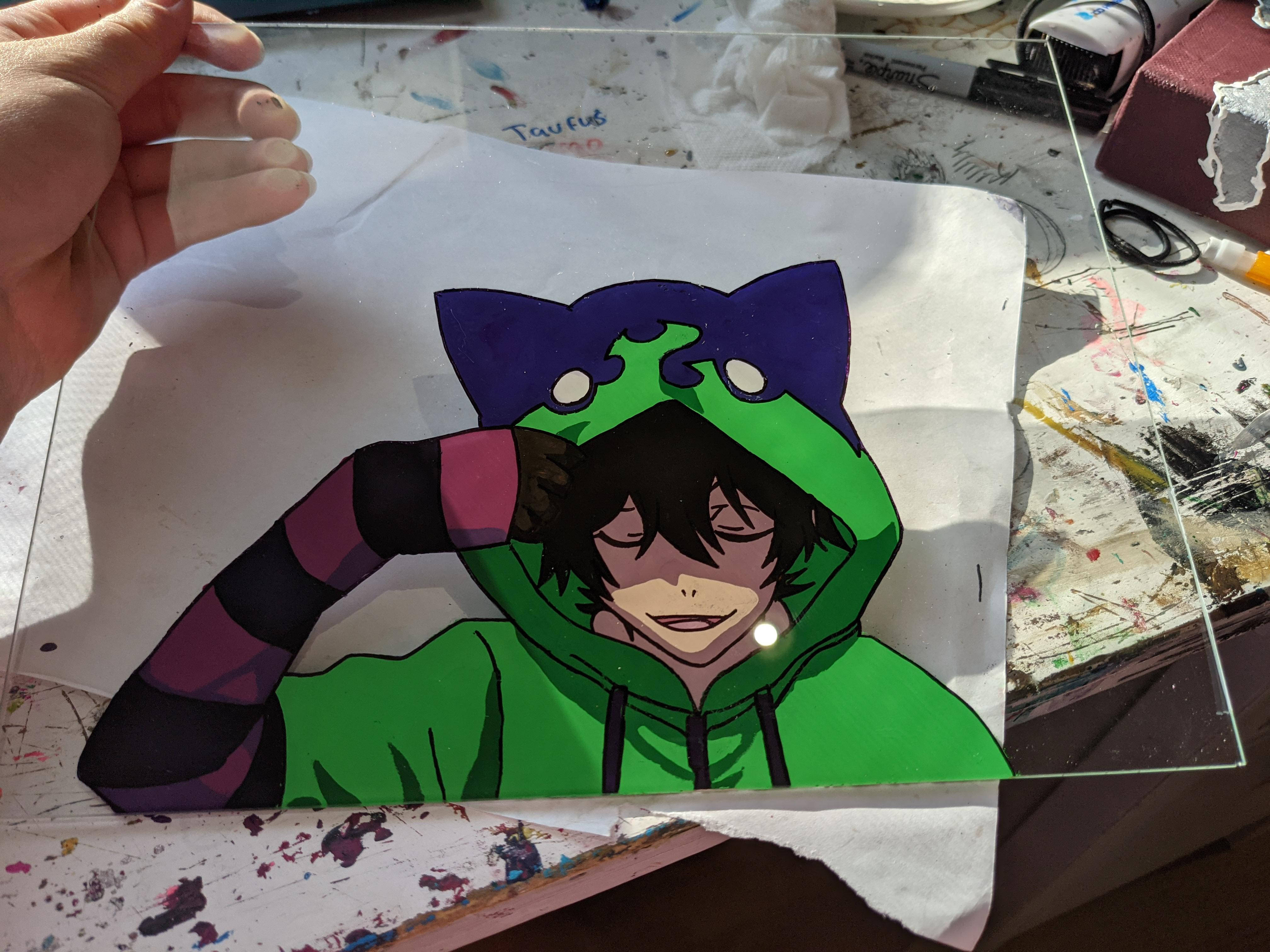 Miya 桌布 壁紙 — 一隻手在濺滿顏料的藝術桌上，拿著一幅色彩繽紛的動漫風格畫作，畫中角色穿著綠色連帽衫，還有紫色貓耳。