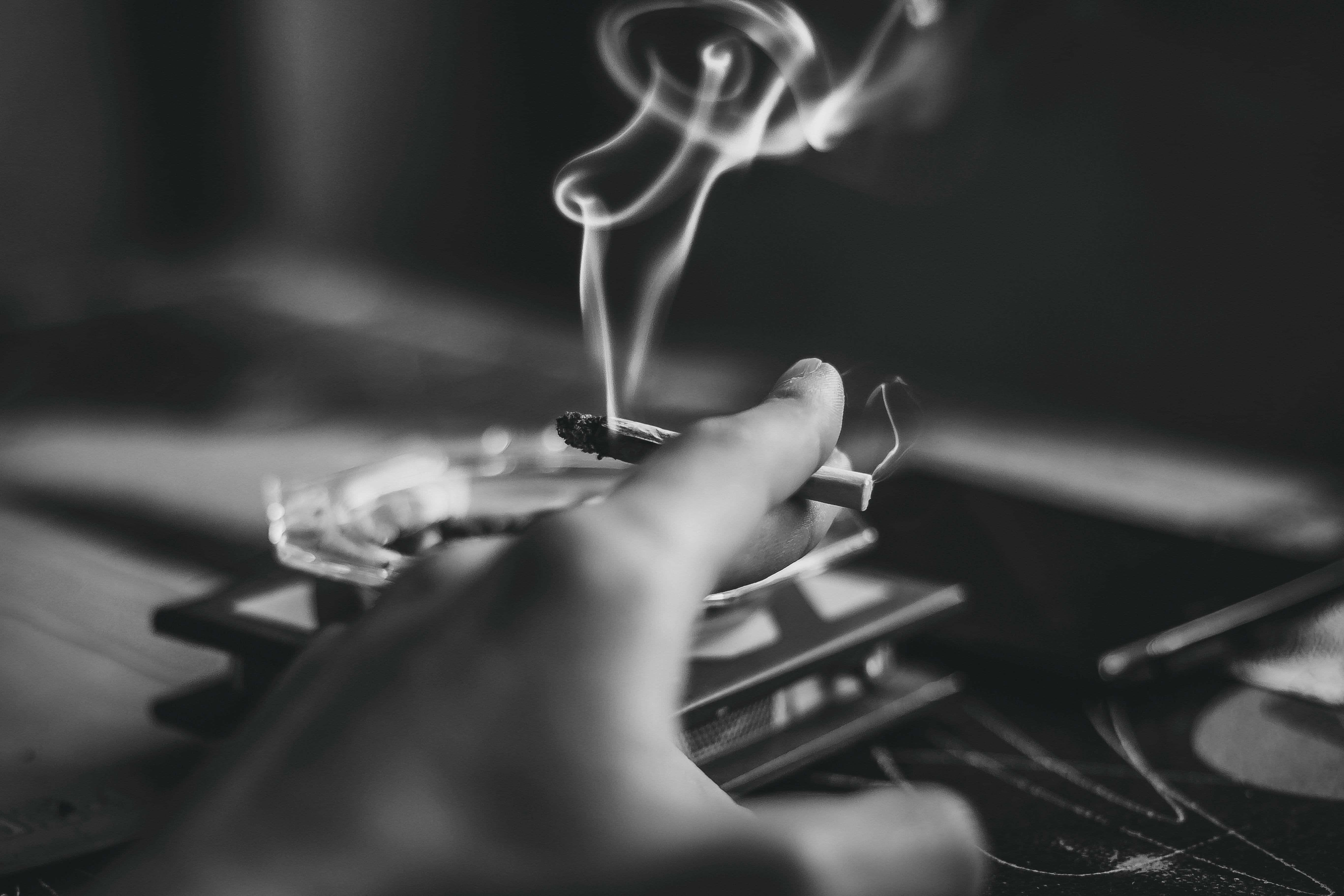 Black Smoke 桌布 壁紙 — 一隻手在煙灰缸上方拿著一支點燃的香煙，煙霧在單色特寫中向上盤旋。