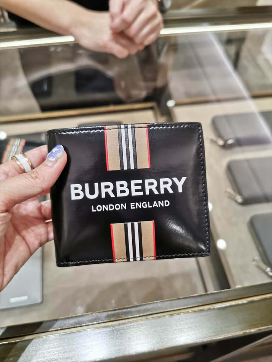 Burberry 桌布 壁紙 — 一隻手在玻璃展示櫃中拿著一個帶有標誌性條紋和粗體白色品牌的黑色 Burberry 皮夾。
