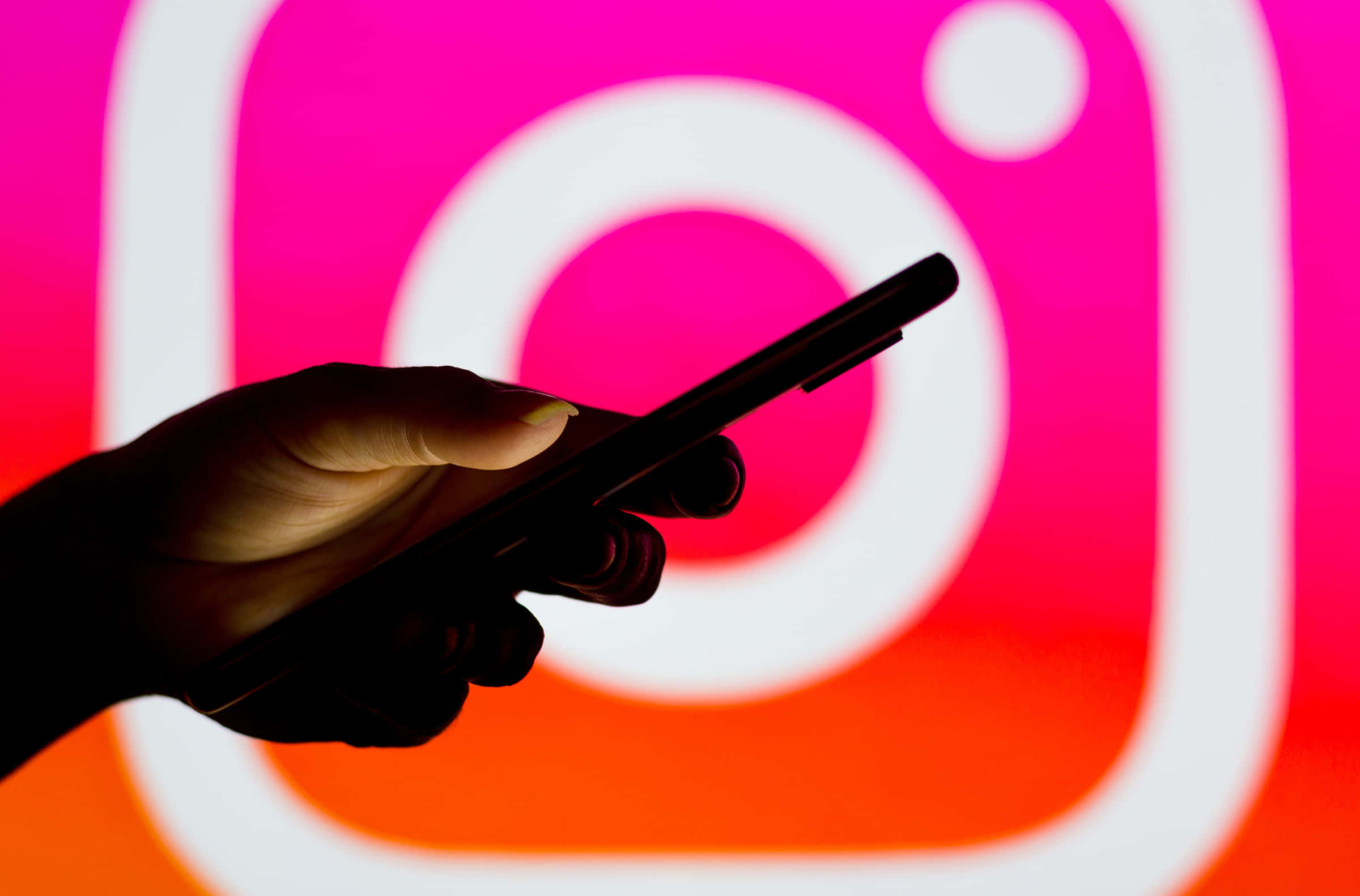 Instagram 桌布 壁紙 — 一隻手在發光的Instagram標誌前拿著智慧型手機，背景是充滿活力的粉紅色和橙色漸變。