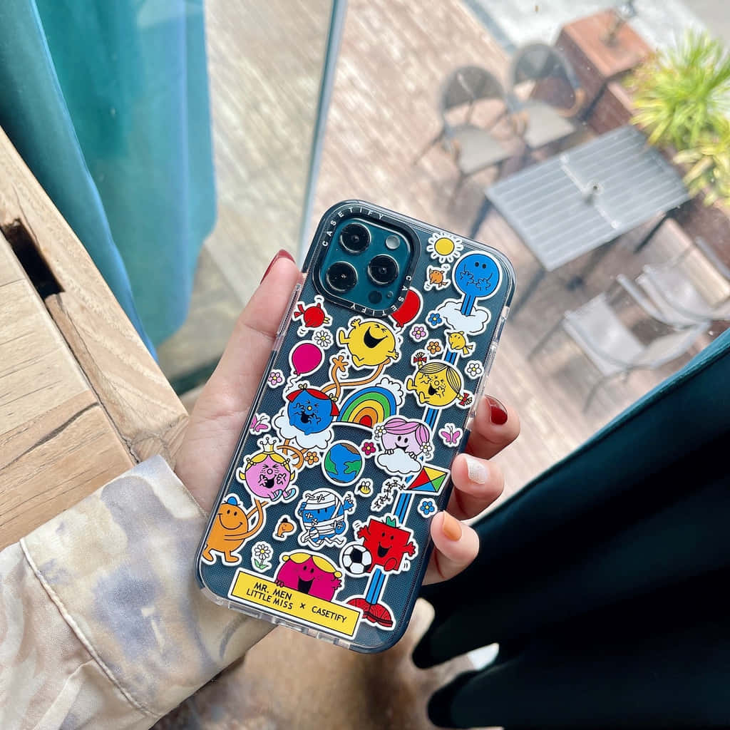 Phone Case 桌布 壁紙 — 一隻手在窗邊拿著一個佈滿色彩繽紛的Mr Men和Little Miss貼紙的手機殼，窗外可見戶外家具。