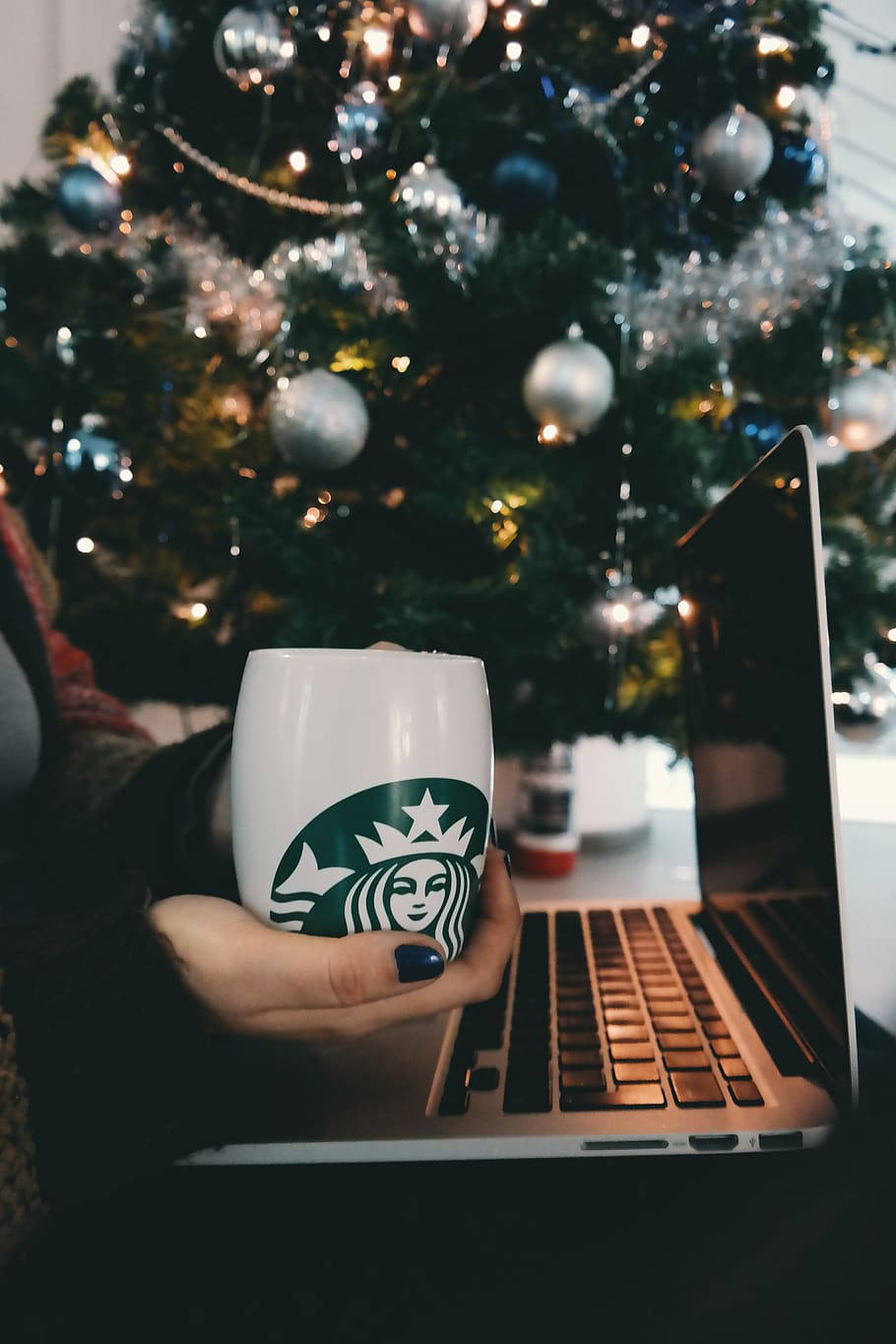 Christmas Tree 桌布 壁紙 — 一隻手在筆記型電腦前拿著星巴克杯子，背景是柔和燈光的聖誕樹。
