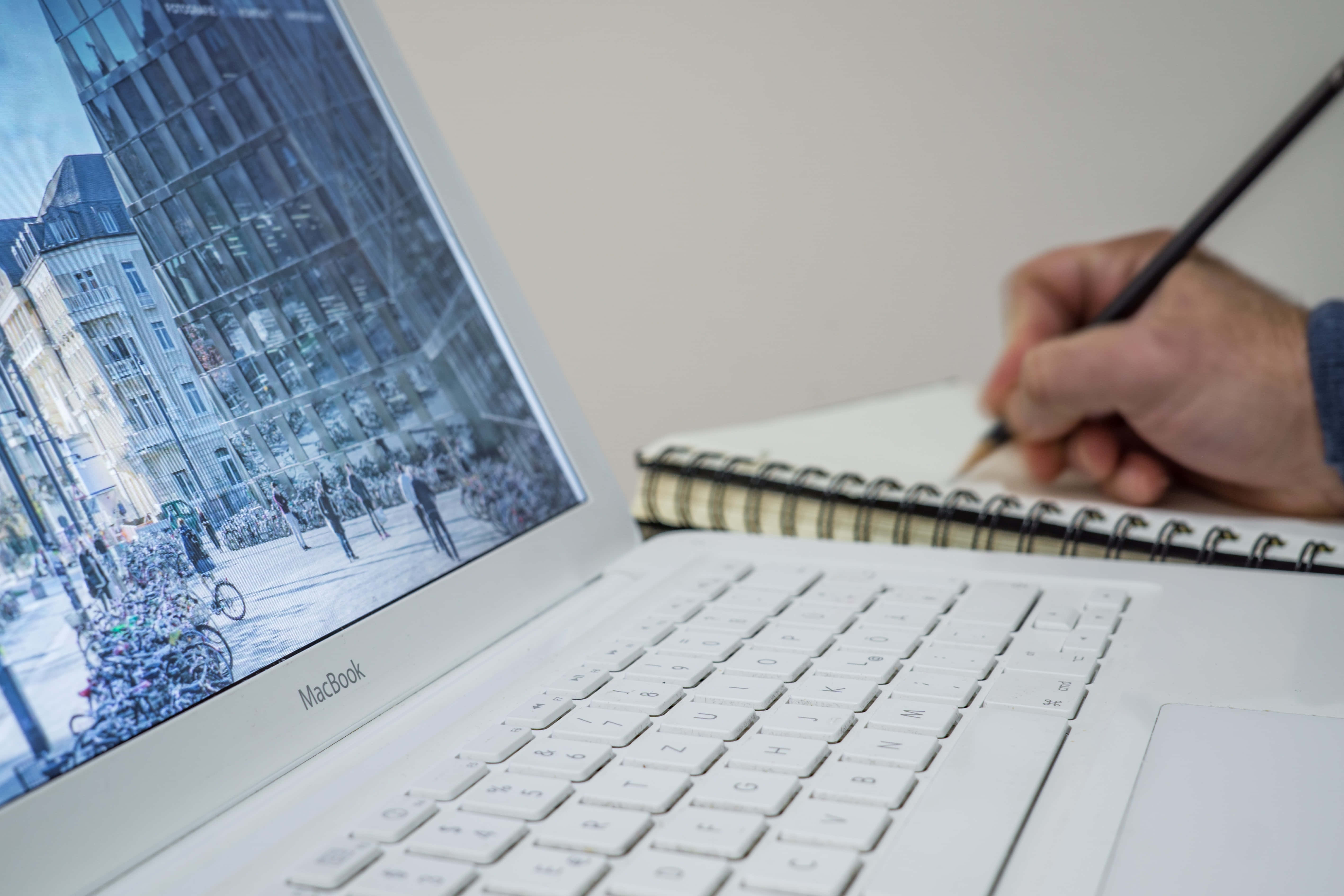 美學 MacBook 壁紙 桌布 壁紙 — 一隻手在筆記本上素描，旁邊的筆記型電腦顯示一個繁忙的歐洲街景，有自行車和歷史建築。