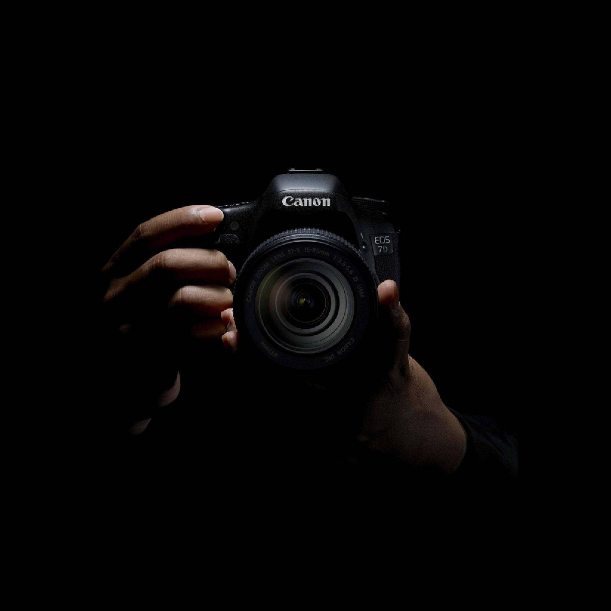 手機 桌布 壁紙 — 一隻手在純黑色背景上以戲劇性的聚光燈拿著Canon EOS 7D DSLR相機。