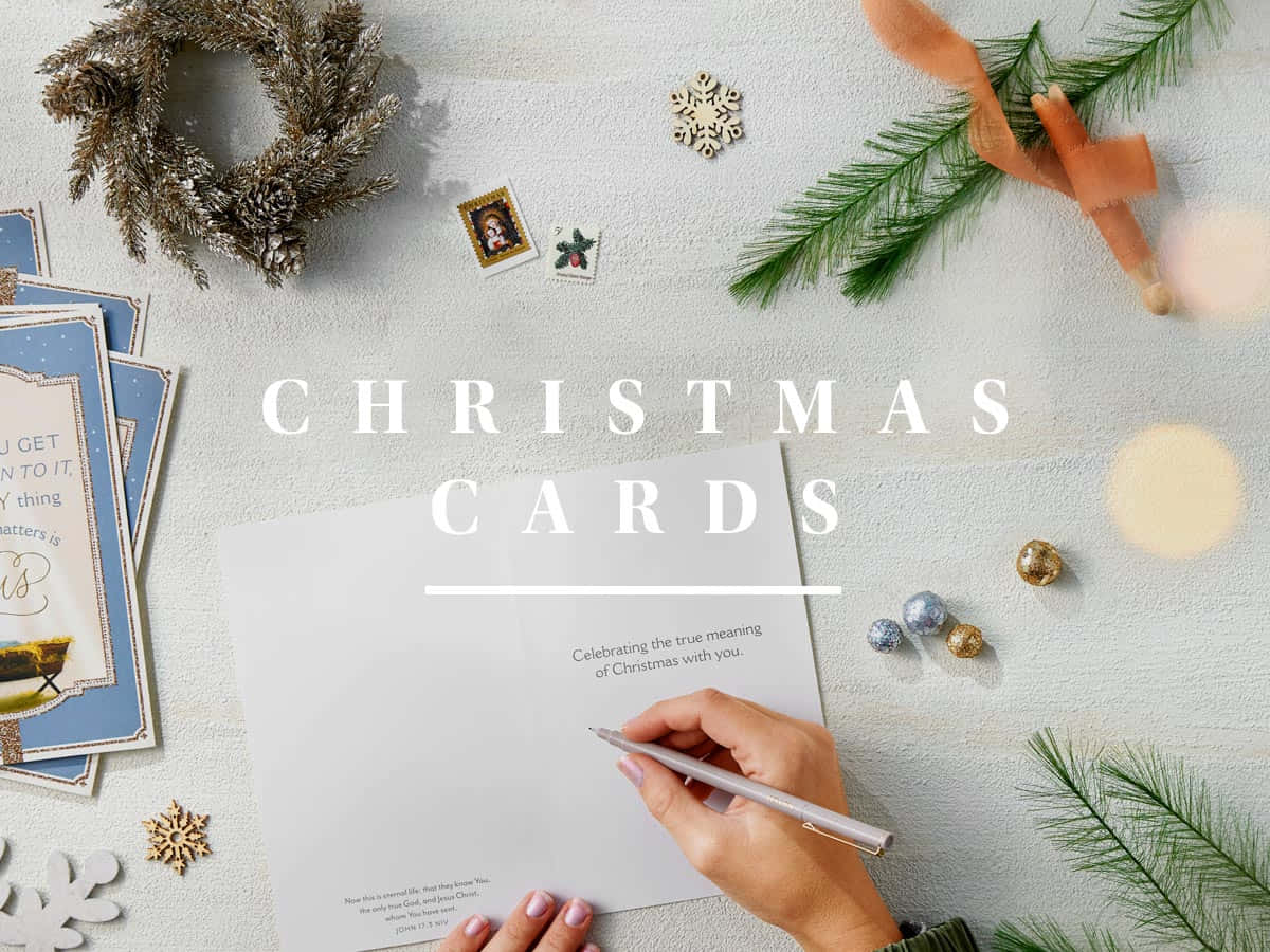 Christmas Card 桌布 壁紙 — 一隻手在聖誕卡上寫字，周圍環繞著松枝、雪花和節日裝飾，位於有紋理的白色表面上。