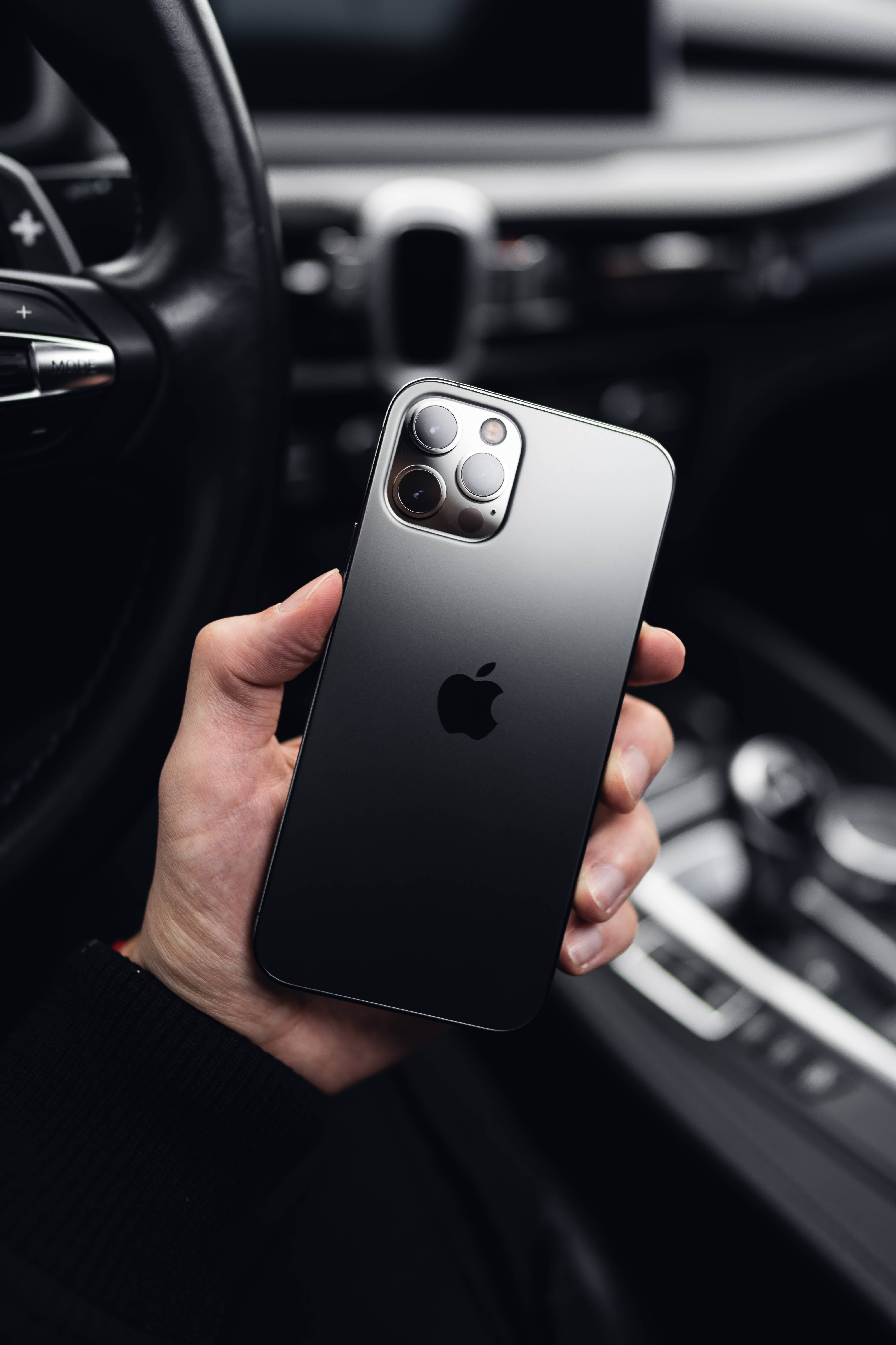 4K iPhone 11 桌布 壁紙 — 一隻手在豪華車內拿著黑色iPhone 12 Pro Max，儀表板和方向盤模糊。
