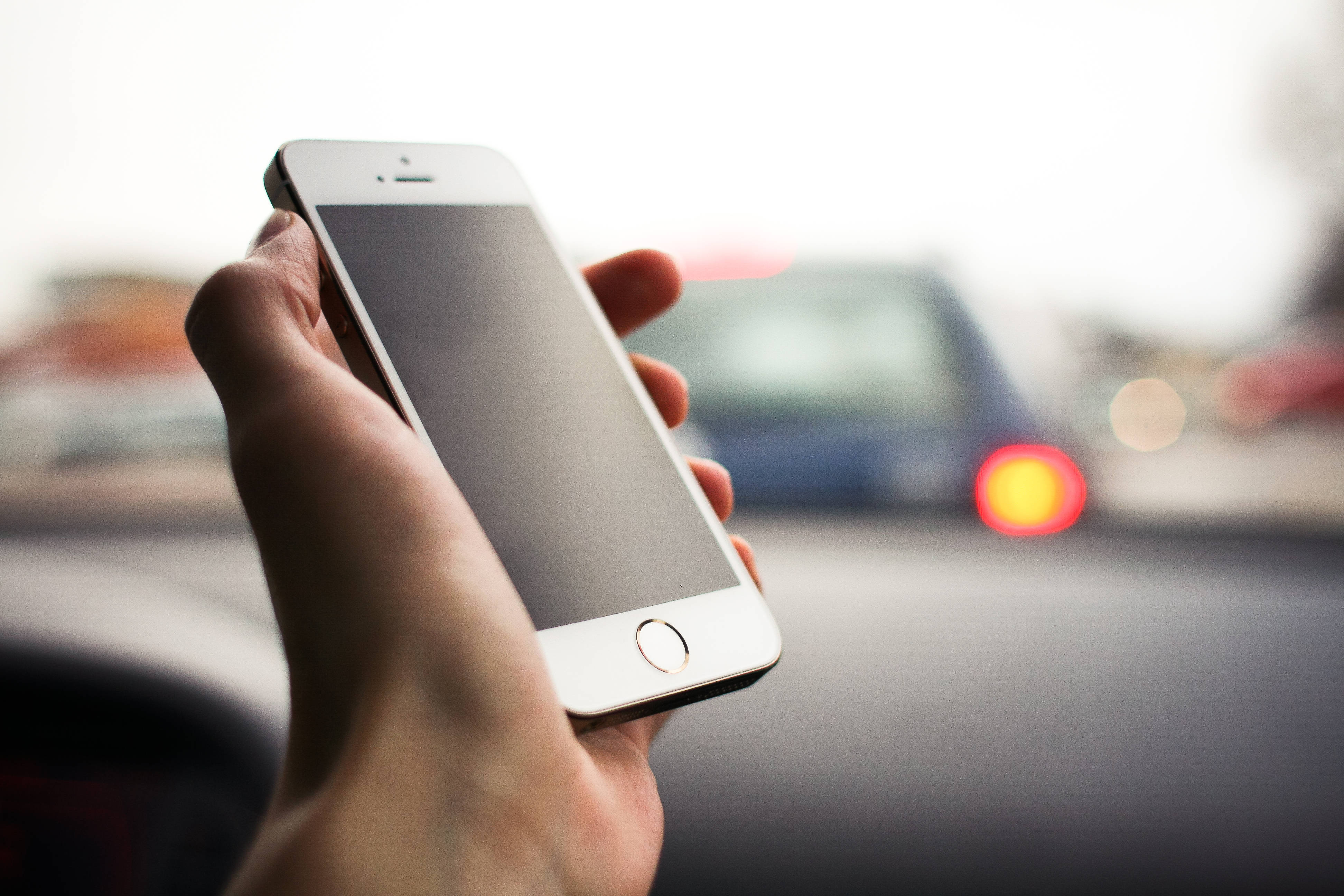 original iPhone 5 桌布 壁紙 — 一隻手在車內拿著一支白色智慧型手機，背景模糊，顯示紅色尾燈和經過的交通。