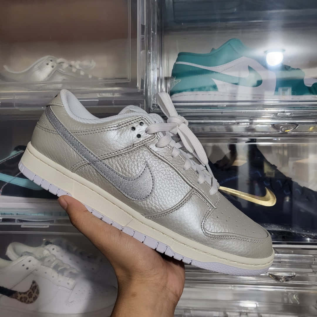 金屬質感 桌布 壁紙 — 一隻手在透明鞋盒前面拿著一雙金屬銀色Nike Dunk Low運動鞋，鞋盒裡裝滿其他色彩繽紛的運動鞋。