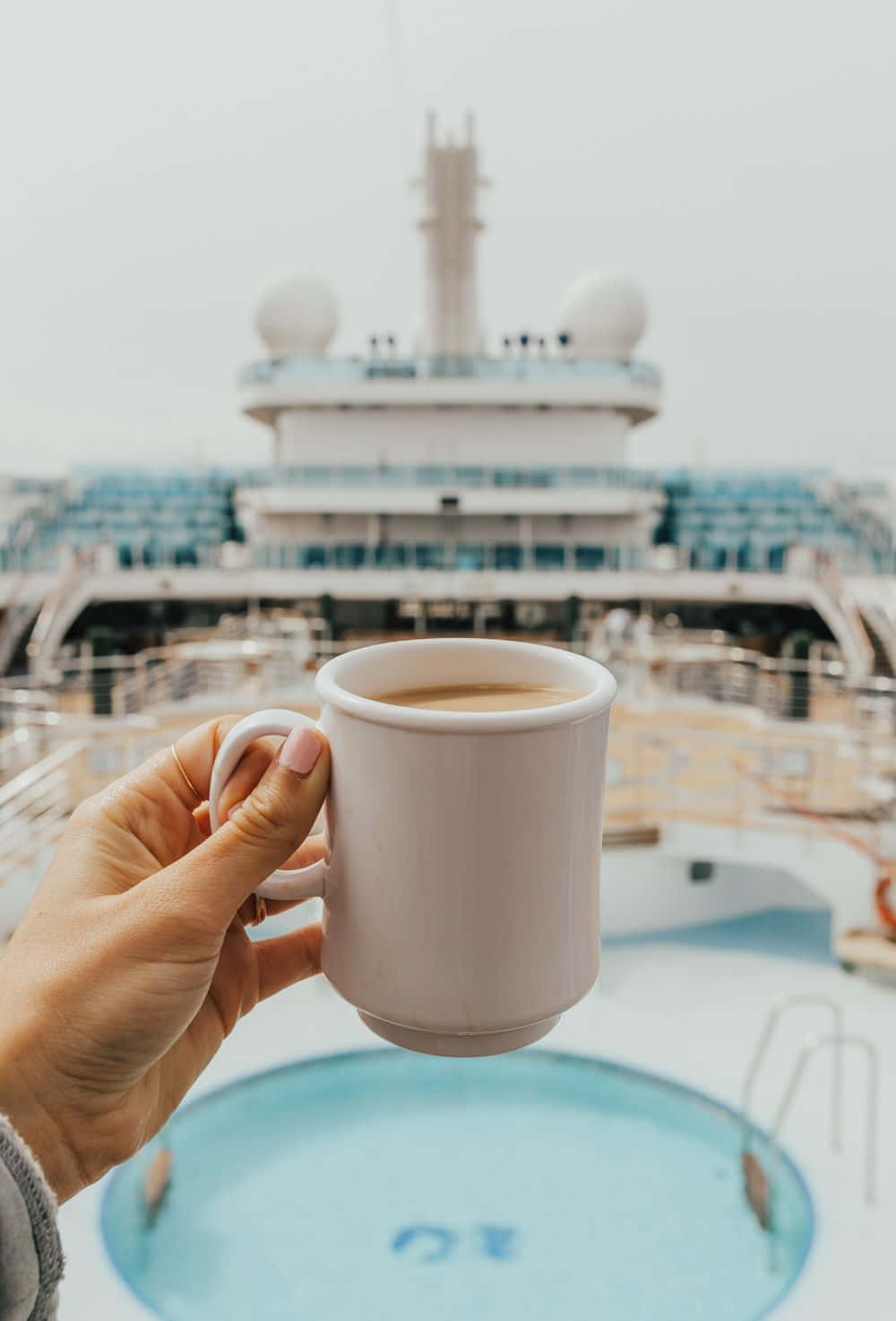 Cruise 桌布 壁紙 — 一隻手在郵輪甲板上拿著一個白色咖啡杯，後面是游泳池和郵輪的上層建築，呈現柔焦效果。