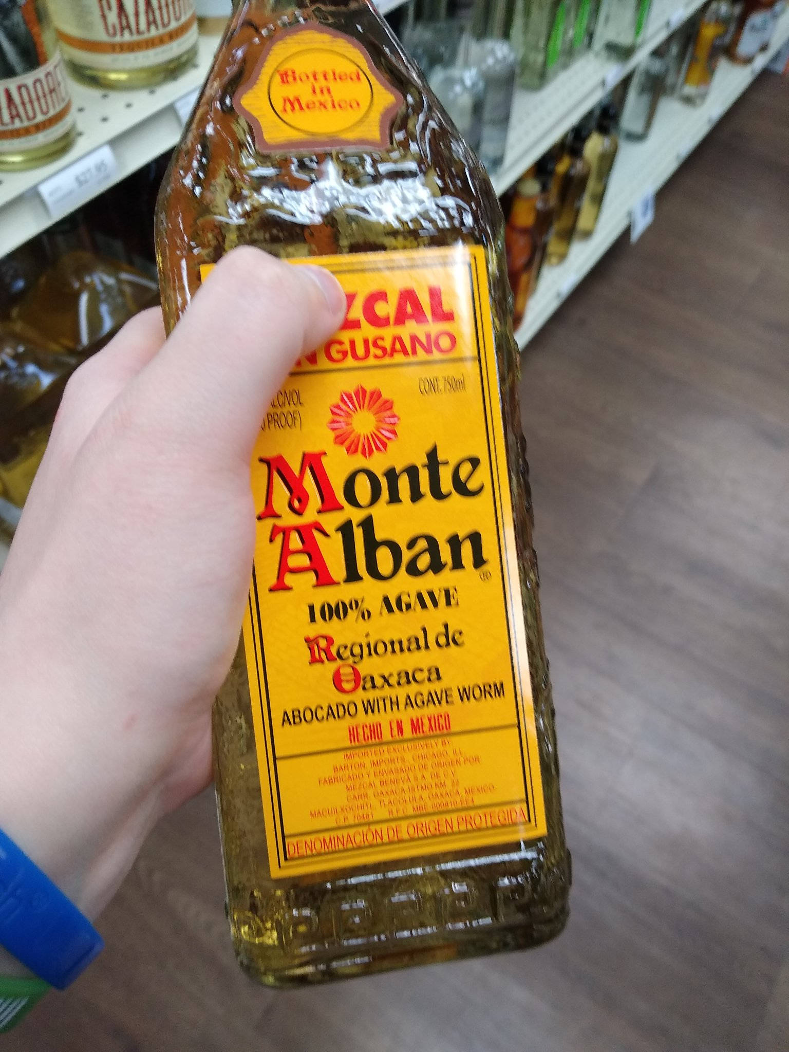 Drink 桌布 壁紙 — 一隻手在酒鋪通道上拿著一瓶帶有亮黃色標籤的Monte Alban龍舌蘭酒。