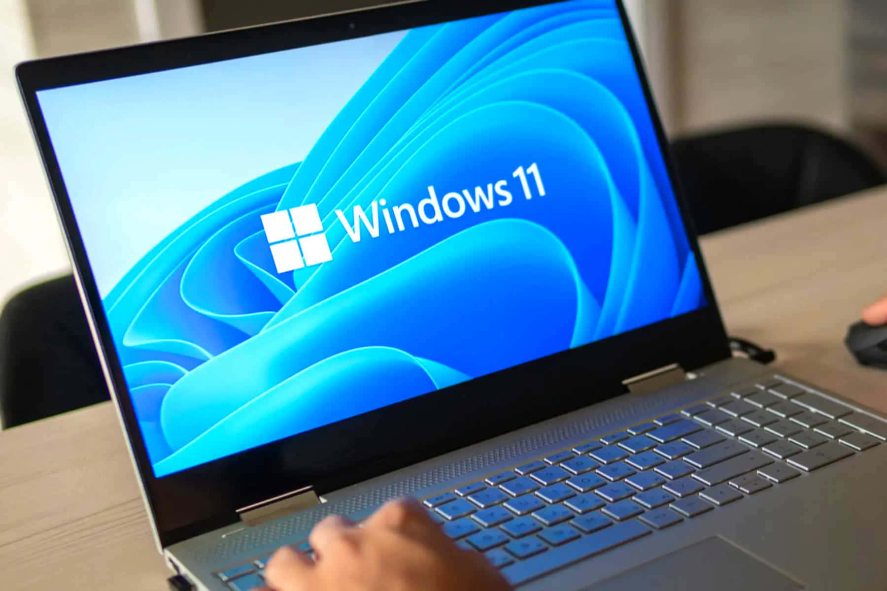 Windows 桌布 壁紙 — 一隻手在銀色筆記型電腦上打字，顯示充滿活力的藍色Windows 11啟動畫面，帶有流動的抽象波浪。