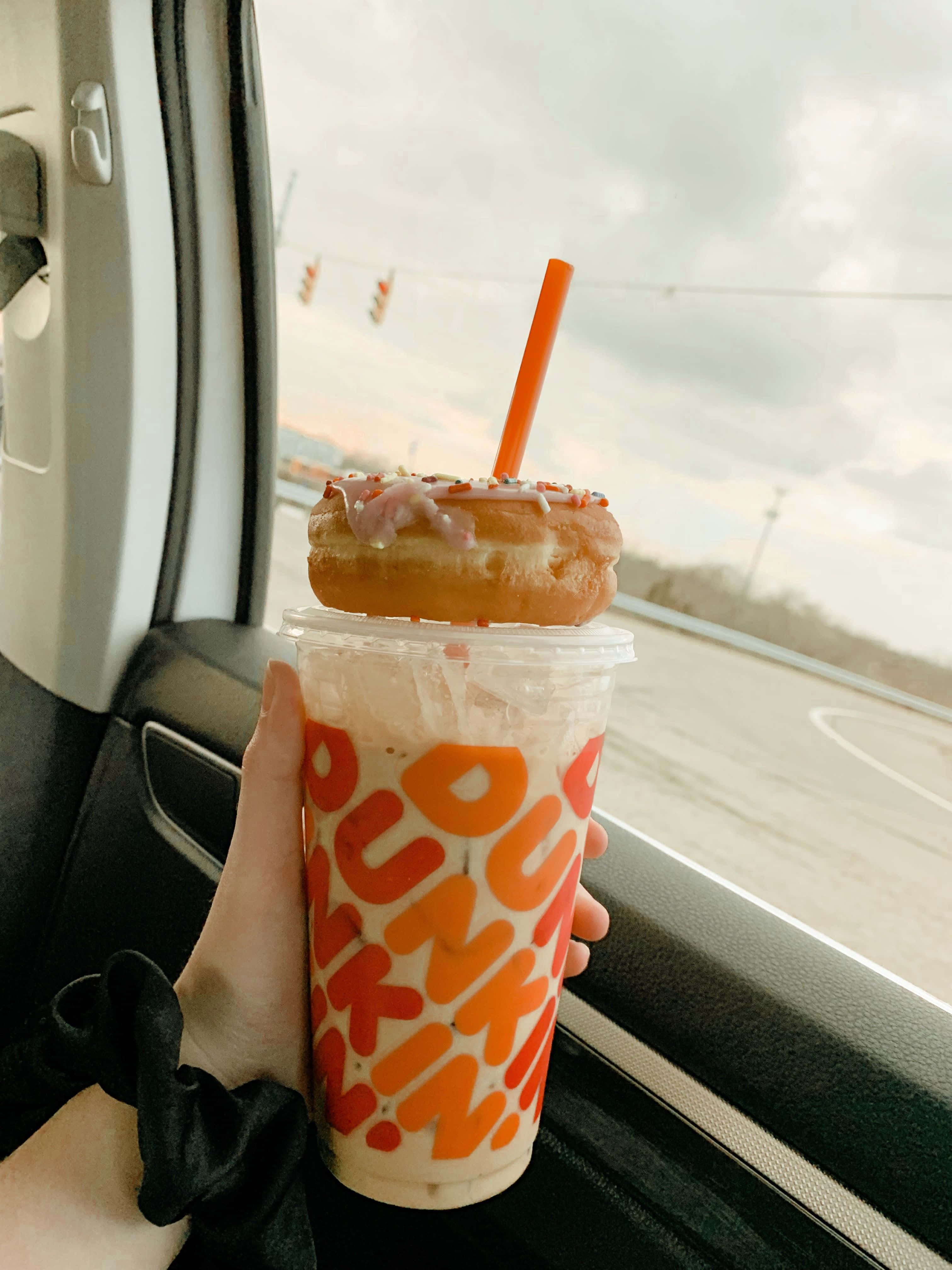 Dunkin Donuts 桌布 壁紙 — 一隻手在陰天透過車窗拿著一杯上面放有糖霜甜甜圈的Dunkin'冰飲。
